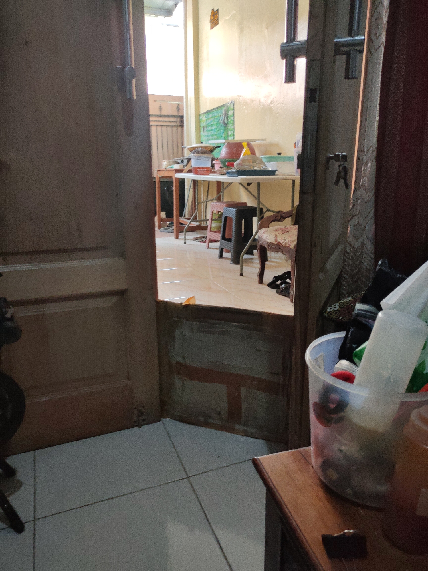 This image shows an interior view of a room, possibly a kitchen or storage area, looking through an open doorway. The foreground is dominated by a wooden door on the left and a wooden table on the right, which holds a clear plastic bucket filled with various items, including plastic bottles and what appear to be cleaning supplies. Beyond the doorway, a cluttered space is visible, featuring a folding table with items on it, a stool, and a chair with floral upholstery. The walls are painted a light yellow, and a patterned curtain hangs on the right side. The flooring appears to be tiled. The scene suggests a functional, lived-in space rather than a pristine one. This location could be a residence in South Jakarta, Indonesia.
