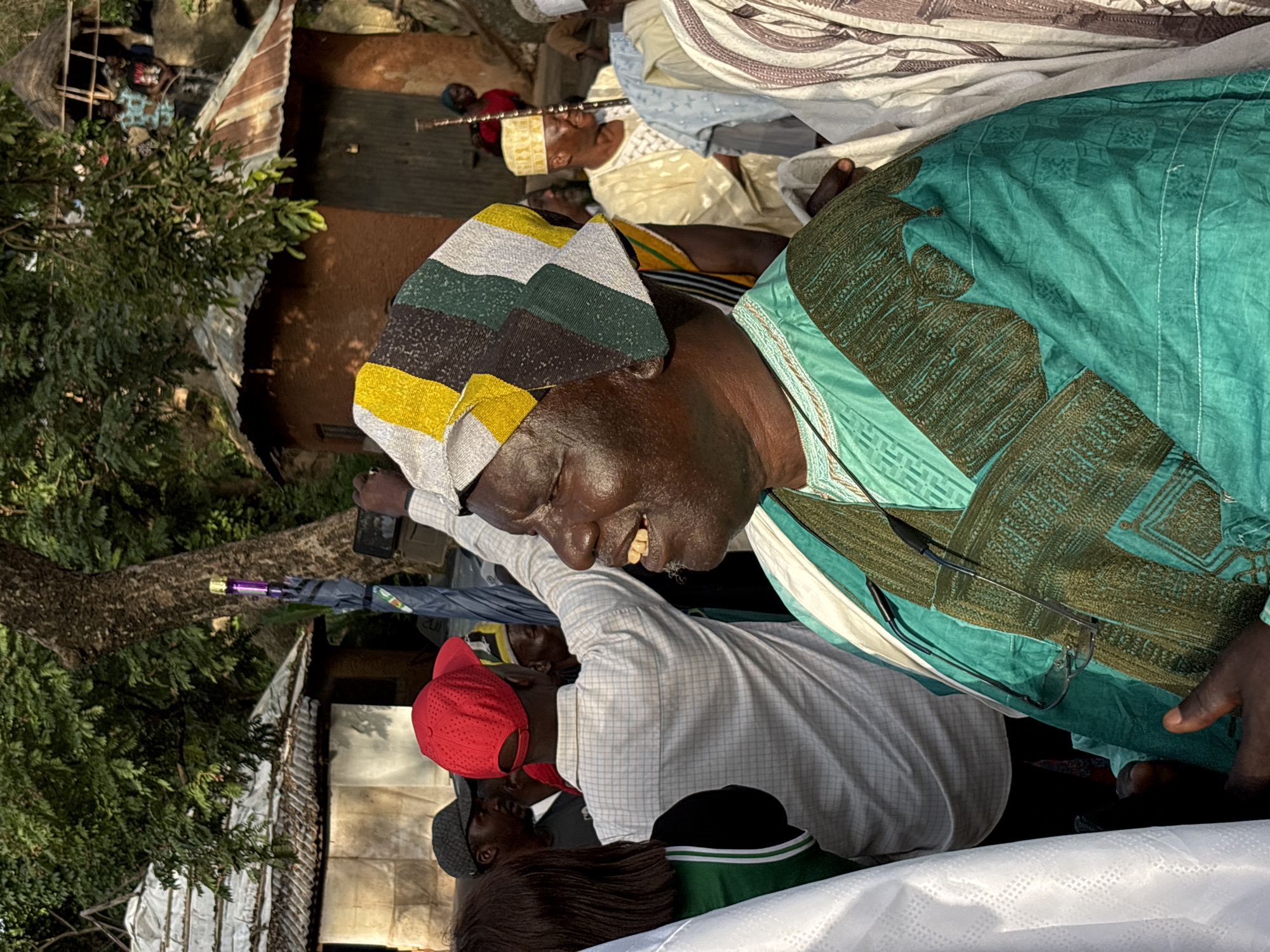 In a dimly lit outdoor scene in Jos, Nigeria, several individuals are gathered. The central focus is an older Black man wearing a green and brown patterned robe and a colorful, striped cap. He is looking down and to the left with his mouth open, revealing his teeth. Next to him, a younger Black man wears a white checkered shirt and a bright red baseball cap, also looking downwards. Partially visible is another person with dark hair wearing a grey cap. The background shows elements of rustic architecture, including a corrugated metal roof and mud-brick walls, partially obscured by lush green trees. A long, thin object, possibly a ceremonial staff or musical instrument, is held by someone in the upper background, who is dressed in a yellow robe and a yellow cap. Another person in the upper background is covered in a patterned white and brown cloth. A purple object, possibly a flashlight, is attached to a tree trunk in the mid-ground. The lighting suggests it is either early morning or late afternoon. The overall atmosphere appears to be one of a gathering or a ceremony.