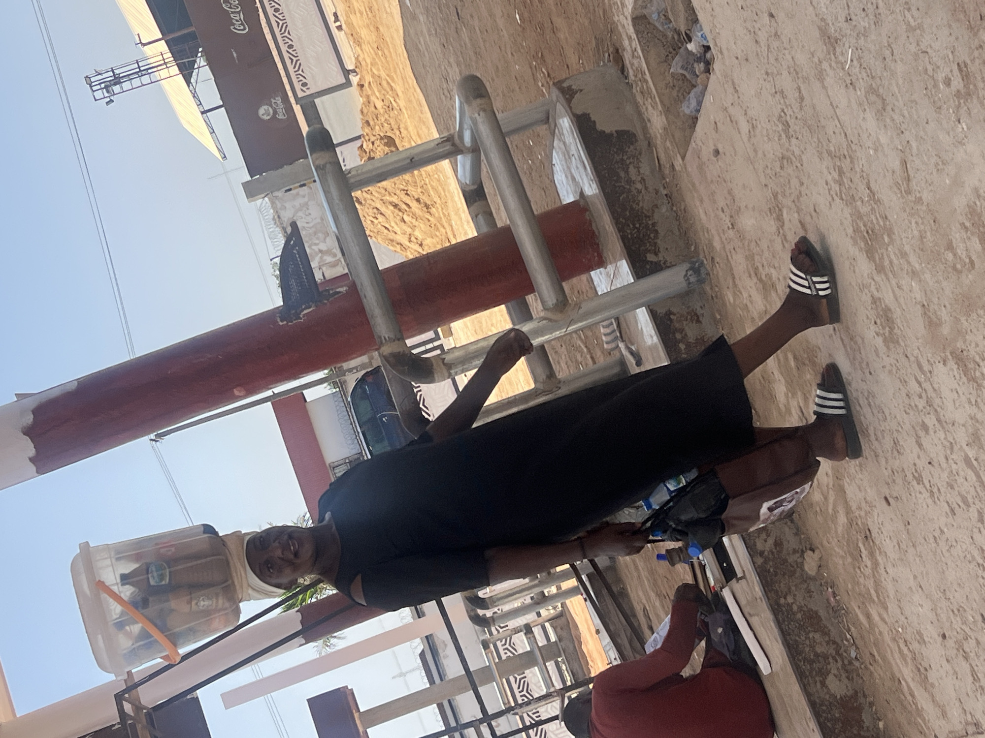 A person wearing a black dress and striped Adidas sandals stands in front of a building structure, likely in Barakin Lerawi, Nigeria. The person holds a motorcycle with a clear plastic bucket filled with assorted items attached to the front. Behind the person, there's a large Coca-Cola sign affixed to the building. A second person is sitting in the lower right of the image, wearing a red shirt. The ground appears to be cracked concrete.