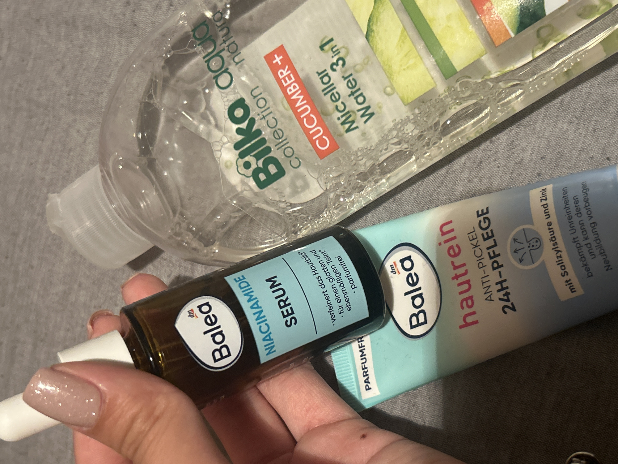 Here is a detailed description of the image:
The image shows three skincare products from the brands Balea and Bilka aqua on a grey background. A person's hand is holding a Balea Niacinamide Serum bottle with a dropper, which is positioned on top of a Balea Hautrein anti-pimple 24H-Pflege tube. Behind these two items and slightly above them, lies a bottle of Bilka aqua Collection Natura Cucumber Micellar Water 3in1. The labels of the skincare products are visible, showcasing the product names and brands.