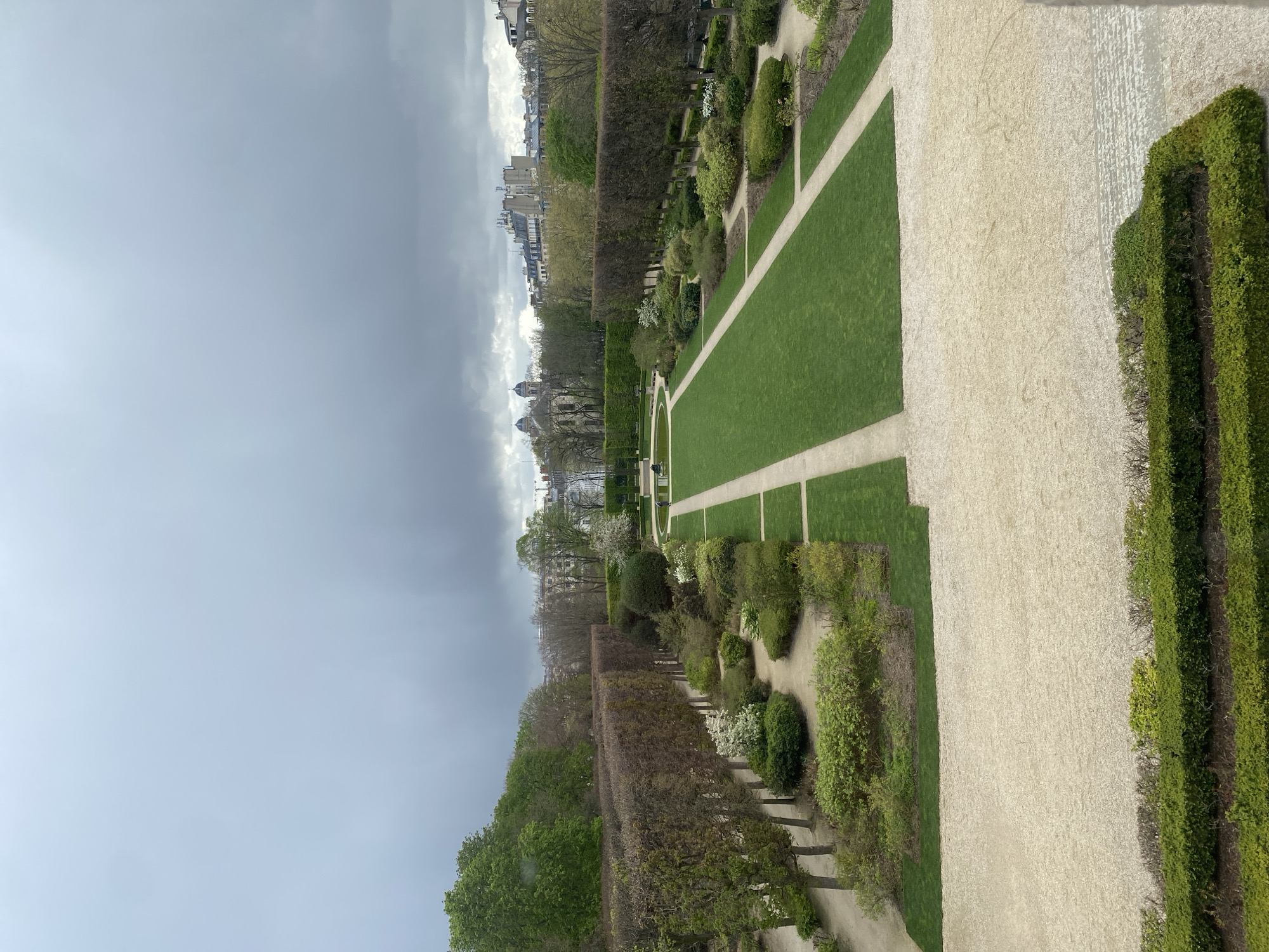 The image captures a detailed, elevated view of the Luxembourg Gardens (Jardin du Luxembourg) in Paris, France, under an overcast sky dominated by heavy grey clouds.
In the immediate foreground, a broad expanse of light-colored gravel paving occupies the bottom third of the frame, bordered by a low, manicured green hedge. Moving into the midground, the garden exhibits a formal layout with meticulously maintained rectangular lawns of vibrant green grass, intersected by light-colored gravel paths. Between these defined areas, various shrub beds contain both green foliage and scattered white blossoms. A prominent feature in the central midground is a circular pond or fountain area, around which a single individual is observed walking on a gravel path, viewed from behind. Several dark-colored park benches are partially visible near this feature.
Further into the background, the garden transitions into sections defined by tall, linear hedges of deciduous trees, some appearing bare or with nascent leaves, while others are densely green. The distant horizon reveals the Parisian urban landscape, characterized by numerous residential buildings with grey rooftops. A notable architectural landmark, the domed structure of the Panthéon, is clearly visible on the left side of the distant skyline, confirming the specific location within Paris. No significant interactions are discernible between subjects.