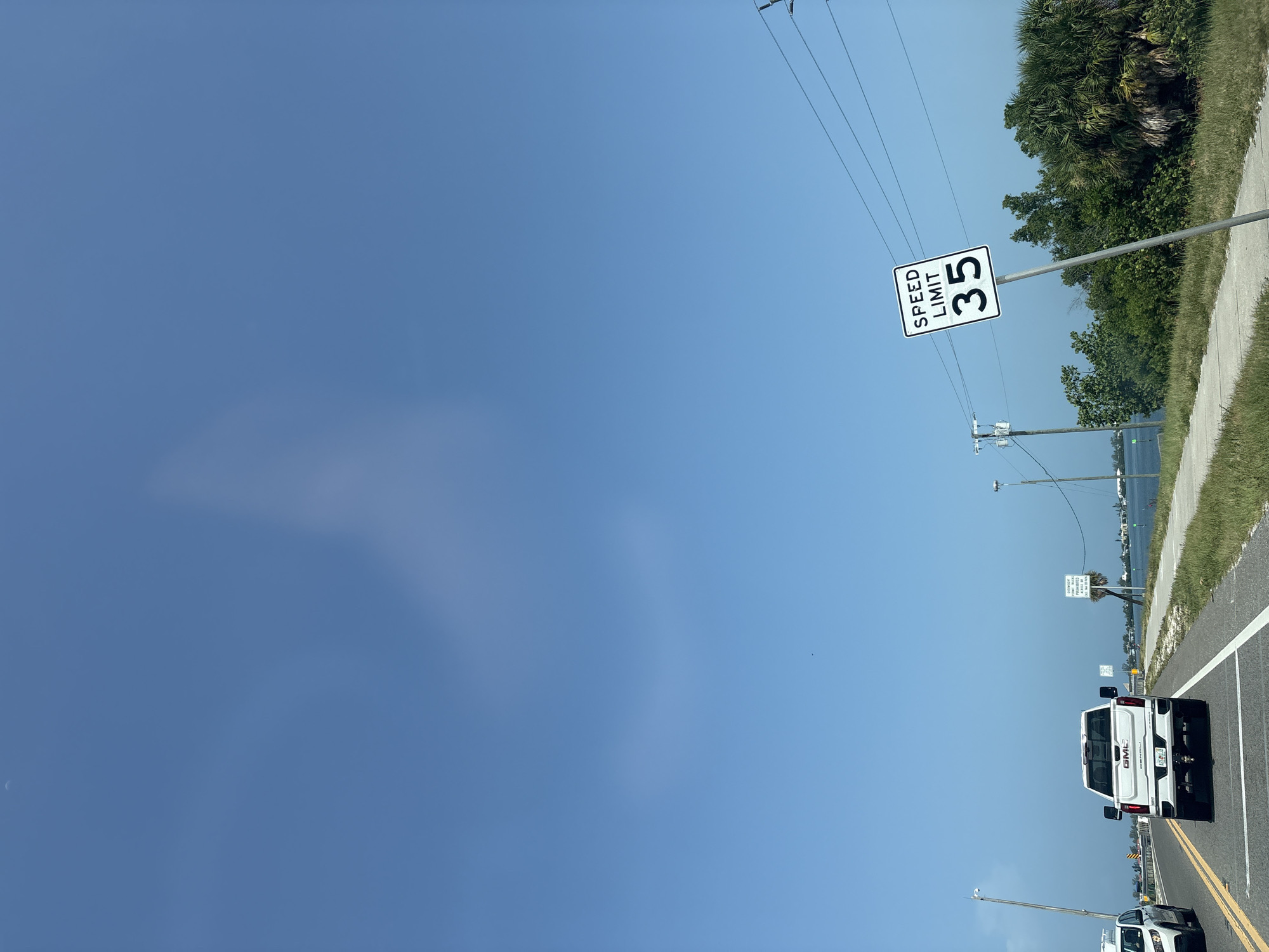 An exterior, eye-level, daytime, medium shot captures a road in Bradenton, Florida, United States of America, bordering a body of water. A white GMC pickup truck is driving away from the viewer on the right side of the road, followed by a white SUV on the left. A speed limit sign indicating "35" is prominently displayed on the right side of the road. The sky is clear blue with faint wispy clouds, and the environment is characterized by lush green vegetation and palm trees. The scene is tranquil, with no visible people or interactions.