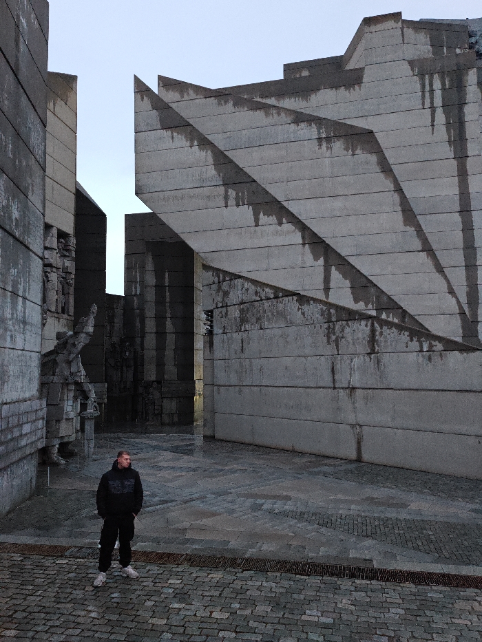The image depicts an adult male standing on wet, patterned paving within the Monument to 1300 Years of Bulgaria in Shumen, Bulgaria. The monument consists of imposing, angular brutalist concrete structures, predominantly grey with dark vertical streaking from water runoff. On the left, several large concrete panels display relief sculptures, while the right features massive, layered concrete slabs forming sharp, ascending geometric angles. The ground is paved with a mixture of grey cobblestones and larger grey flagstones, showing wet surfaces. A metal grate is visible in the foreground. The male, positioned slightly left of center, wears a black hooded sweatshirt, black athletic pants, and white athletic shoes. He stands with his hands in his pockets, looking towards the left. The sky is overcast and light grey. No other individuals are present.