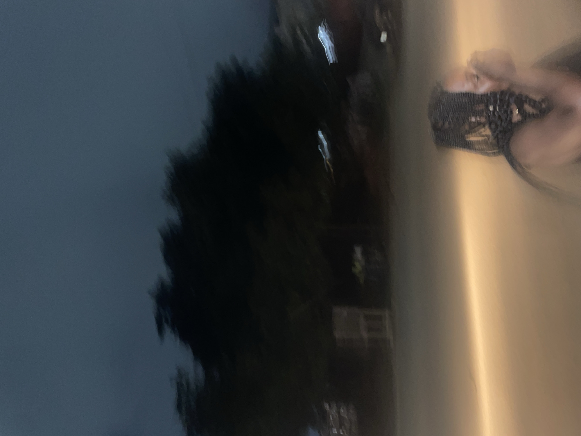 An out-of-focus image depicts a dark, hazy outdoor scene, likely at night. The sky is a deep, muted blue, with indistinct dark shapes of trees occupying the upper middle portion of the frame. Below the trees, the environment is illuminated by a bright, diffused light source, casting a warm, orange glow across the lower half of the image. In the foreground, the blurry figure of a person with braided hair is visible, their hands raised to their face. The scene suggests a location in Guzape, Nigeria, possibly a street or an open area, given the low light and the presence of what appear to be streetlights or headlights contributing to the overall illumination.