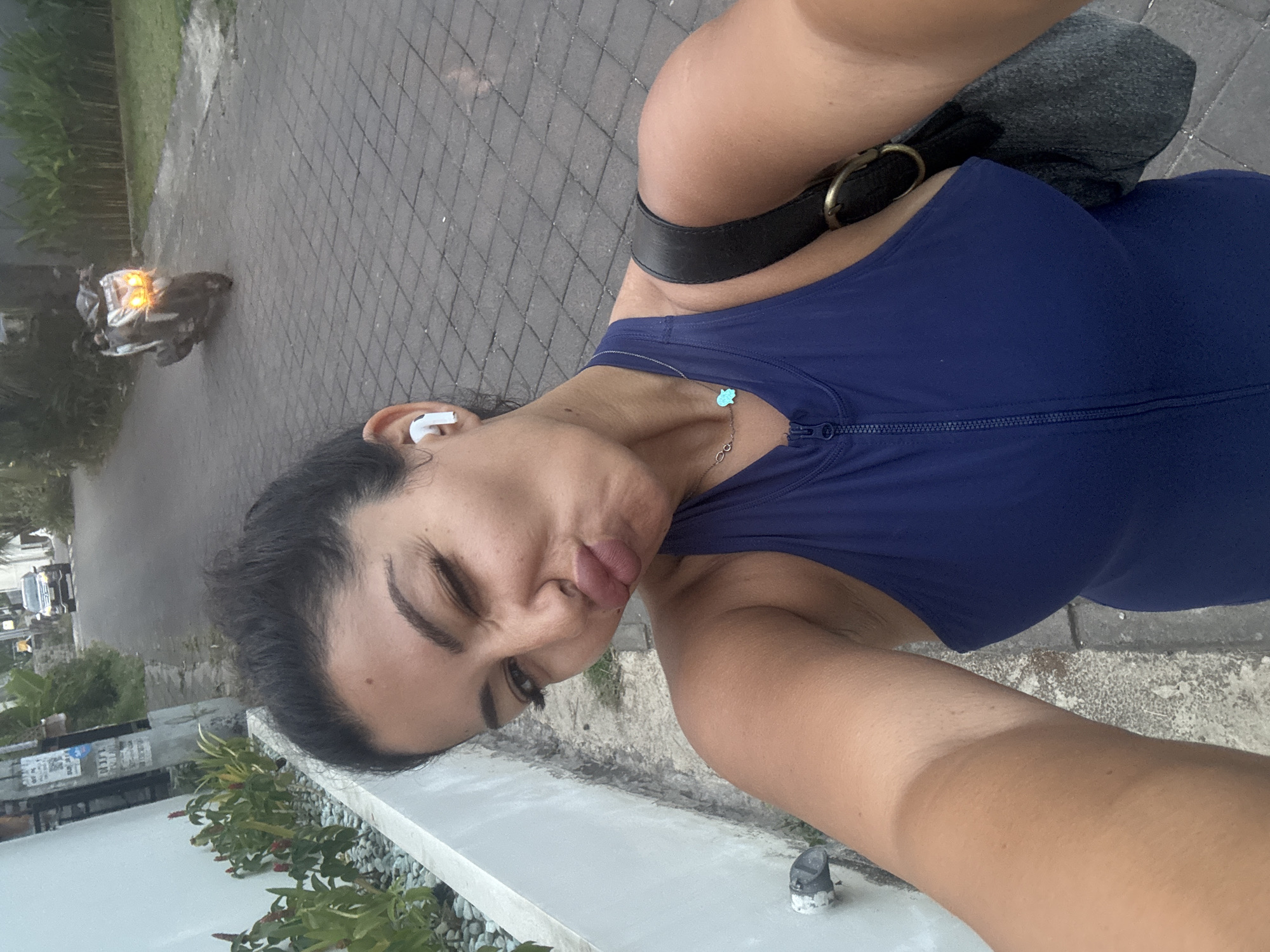 In Kuta Selatan, Indonesia, a woman takes a selfie outdoors. She is standing on a paved walkway. She is wearing a dark blue, sleeveless zip-up top and a delicate necklace with a blue pendant. She has white earbuds in her ears and is winking at the camera with her lips pursed. She has a dark bag slung over her right shoulder. In the background, a scooter with illuminated headlights drives down a road. A car is visible further down the road. Lush green plants line the left side of the walkway, and a white wall is adjacent to the plants. The overall lighting suggests it is late afternoon or early evening.