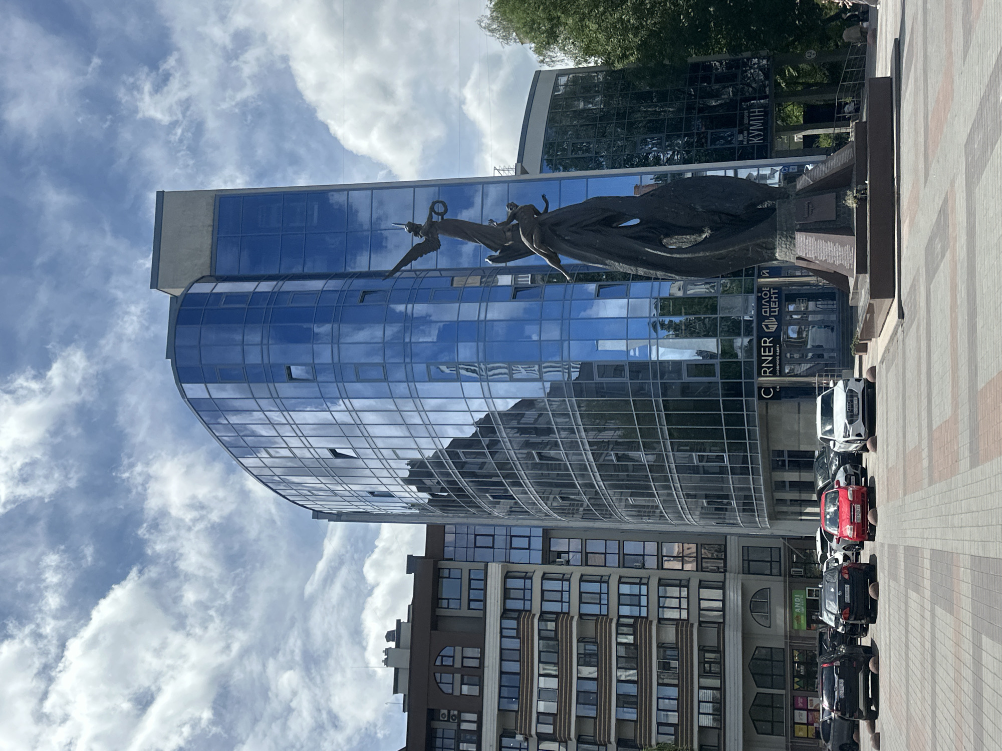 The image depicts an outdoor scene in Ternopil, Ukraine, featuring a modern office building with a curved glass facade that reflects the sky and surrounding structures. In the foreground, a dark bronze sculpture stands on a tiered stone base. The sculpture appears to represent two human figures, one appearing to be a winged female figure holding a wreath, reaching down to another figure. The base of the sculpture has inscriptions and a small floral arrangement.
To the left of the main building is a multi-story residential building with a more traditional architectural style, characterized by balconies and numerous windows. In front of these buildings is a paved public square with a parking area. Several cars are parked in the square, including a red Fiat, a black Audi, and a white Audi. The sky above is partly cloudy with dramatic cloud formations against a blue background.
The ground of the square is paved with square tiles arranged in a patterned walkway. To the right of the main building, trees and part of another building are visible. Signs on the buildings indicate businesses and services, with some text in Ukrainian. The overall scene suggests a well-maintained urban public space.