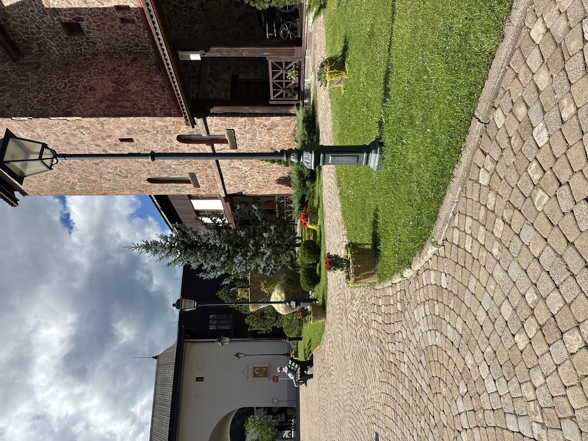 The image depicts a courtyard area within Estosadok, Russia. The scene features a cobblestone pathway curving through a well-maintained lawn. A solitary individual, dressed in a light-colored hat and jacket, sits on a dark bench, facing away from the camera. To the right of the pathway stands a tall, ornate lamppost. Behind the lamppost, a large, textured stone building with arched windows and a wooden-shingled roof dominates the right side of the frame. Various potted plants and green shrubs are strategically placed throughout the courtyard, adding to the manicured appearance. In the background, a large archway leads to another building, and more trees and foliage are visible. The sky above is partially cloudy, with patches of blue sky.