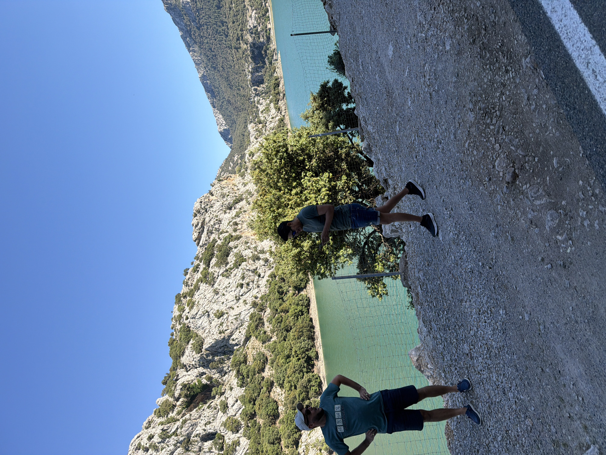 The image shows a landscape in Escorca, Spain, featuring two men in shorts and t-shirts standing on a gravelly roadside overlooking a lake or reservoir surrounded by mountains. The sky is clear blue. One man is wearing a white baseball cap. A fence runs along the edge of the road, separating it from a steep drop to the water.