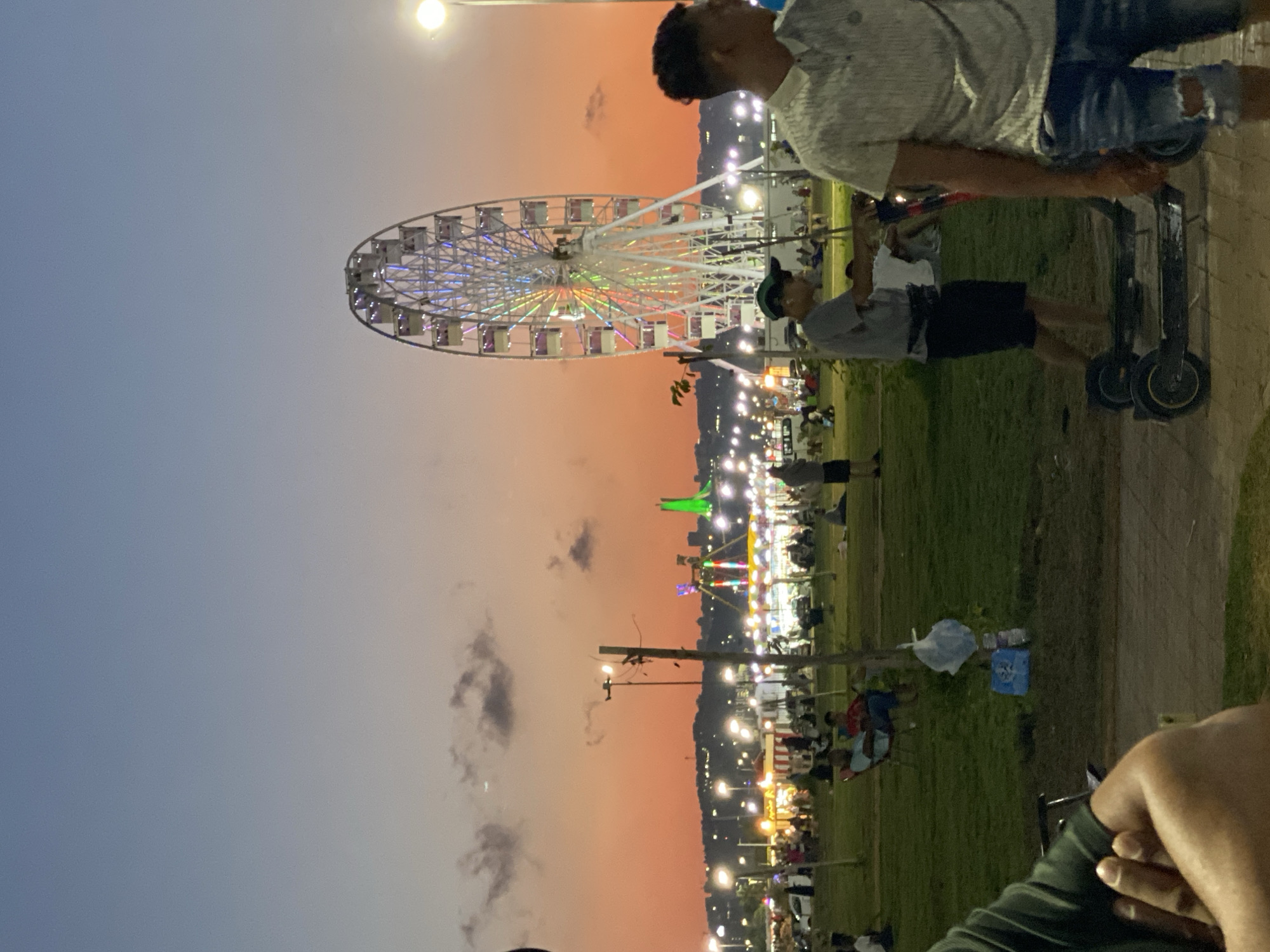 The image depicts a public park at dusk, with a Ferris wheel illuminated in the background. The sky is a gradient of orange and blue, indicating the transition from sunset to twilight. Several people are visible in the park, some sitting on benches or the grass, while others are standing or walking. Two individuals are shown riding electric scooters in the foreground. In the distance, beyond the park, a city skyline is visible with numerous lights. The scene suggests a recreational area with amusement park attractions, likely within or near the city of Hussein Dey, Algeria, given the presence of typical city structures and infrastructure. The overall atmosphere is relaxed and lively, with people enjoying an evening out.