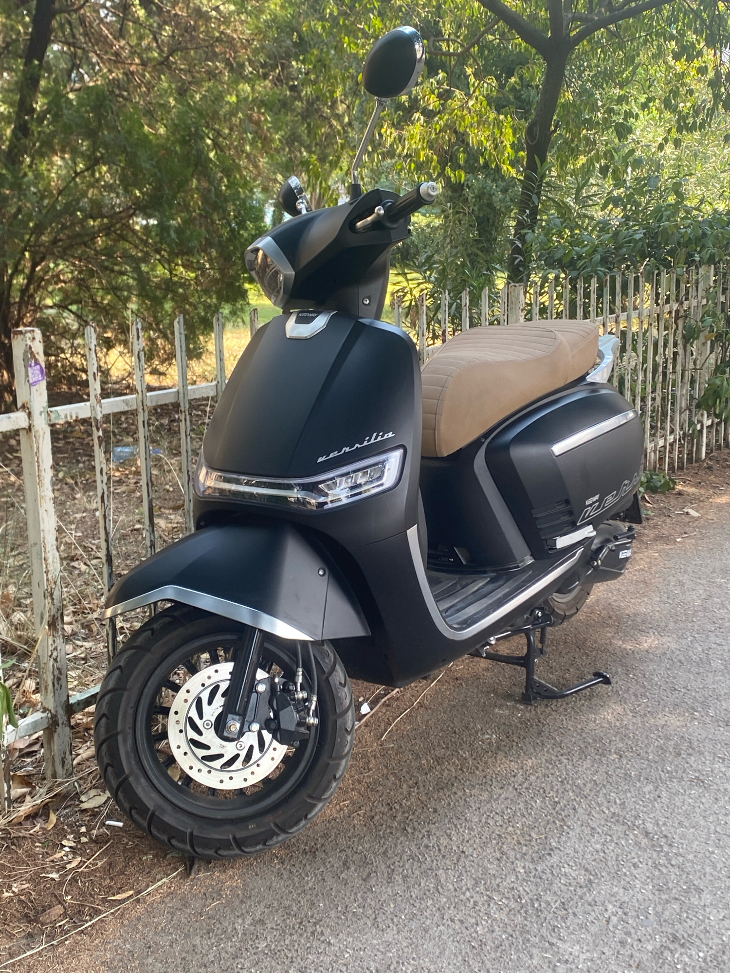 A black scooter with a tan seat is parked on an asphalt road next to a white metal fence. The scooter is angled towards the viewer, with its front wheel turned slightly to the left. The scooter has a sleek, modern design with a matte black finish and chrome accents. The seat is a light tan color and appears to be made of leather or a similar material. The fence is made of white metal bars and is partially covered in ivy. Behind the fence, there are trees with green leaves. The ground is covered in dry leaves and twigs. The scene is lit by natural sunlight, casting shadows on the ground. It is difficult to determine the exact location in Podgorica, Montenegro, as there are no specific landmarks visible in the image.