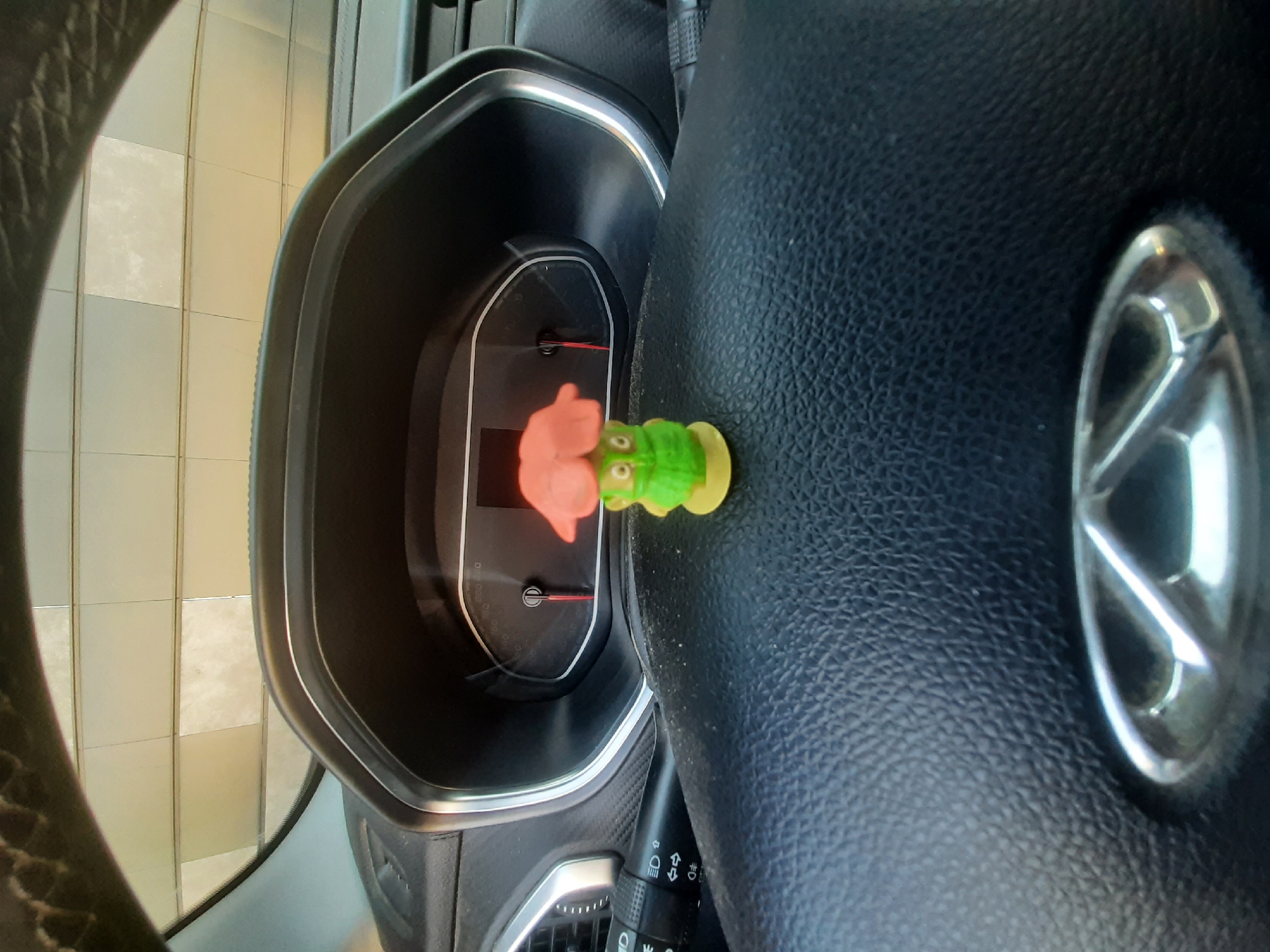The image shows a close-up view of a car's interior, specifically the dashboard and steering wheel. A small, green, frog-shaped toy is prominently placed on the steering wheel. The dashboard features a digital display with a red needle, suggesting it's part of the instrument cluster. The steering wheel is black and textured, with a silver emblem in the center. To the left of the dashboard, a tiled wall is visible through the car window, with square tiles in varying shades of beige and white, possibly indicating an indoor setting like a garage or a car wash. The lighting appears to be artificial or overcast, as there are no strong shadows, and the overall tone is somewhat muted. The image is taken from a slightly tilted angle, giving a dynamic perspective of the car's interior. There is no visible text in the image. The activity taking place is not clear, but the presence of the toy suggests it might be a personal vehicle with decorative items.