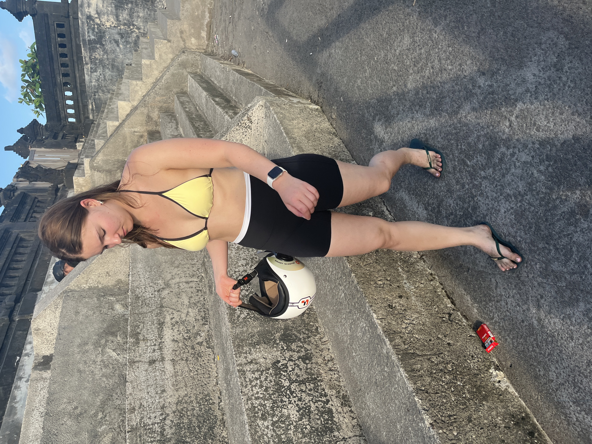 A young woman stands on concrete steps, holding a white helmet in her right hand. She is wearing a yellow bikini top, black shorts, and dark green flip-flops. Her left wrist is adorned with a smartwatch. To her left, a red pack of cigarettes lies on the ground. The background shows a stone structure and a clear sky. The scene appears to be an outdoor public space in North Kuta, Indonesia.