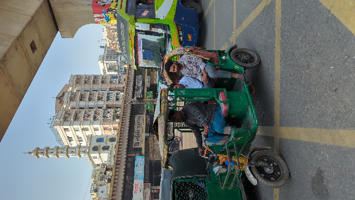 An auto rickshaw is parked on a street with yellow lane markings. Two passengers are seated in the back, while the driver sits in the front. Buildings and a tall tower are visible in the background. A large bus is partially visible next to the auto rickshaw. An overpass is situated above the street.