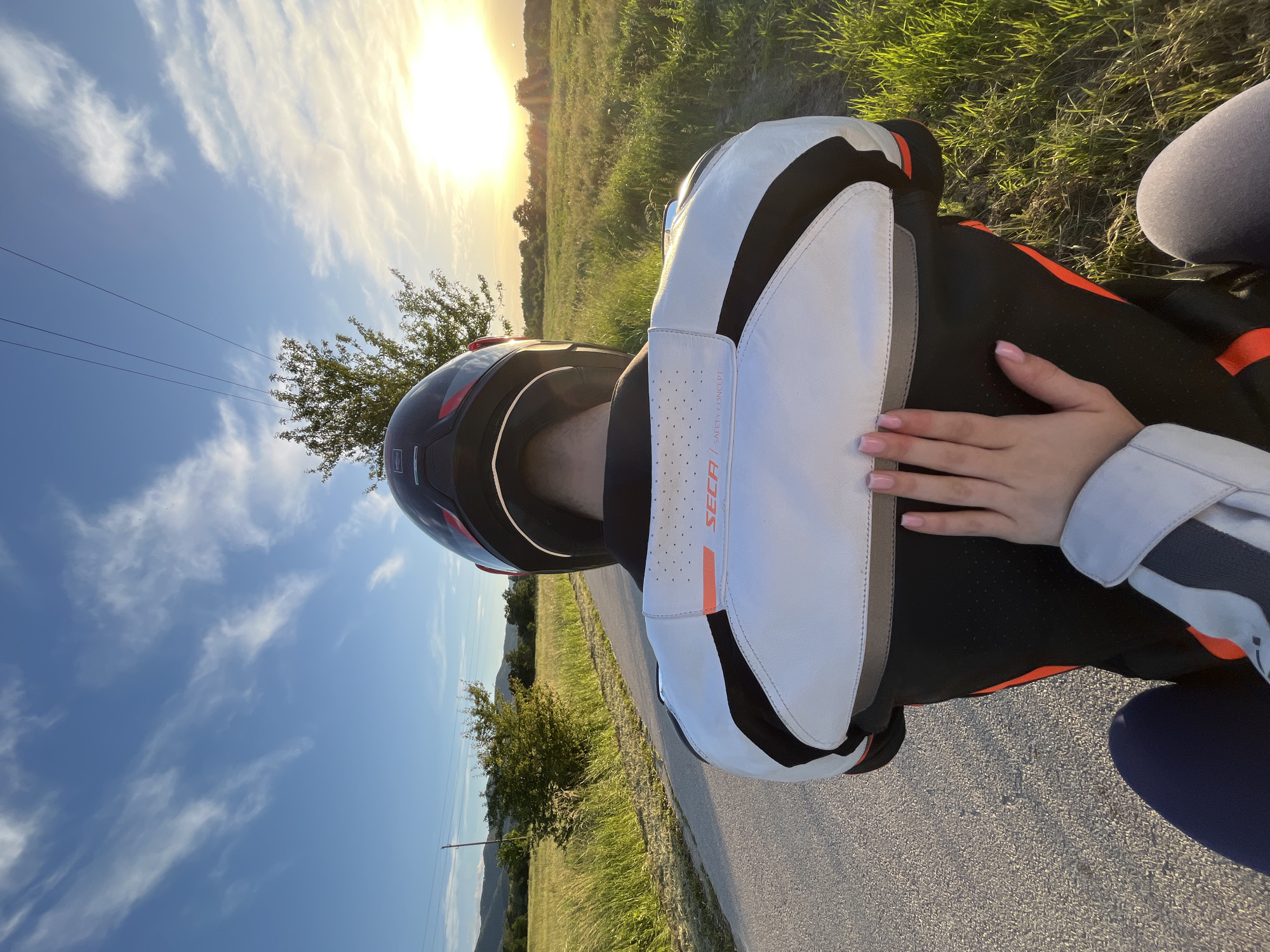 A person wearing a motorcycle helmet and protective gear is sitting on a motorcycle on an asphalt road. The person's hand rests on their lap. The background features a field with green grass, trees, and a bright setting sun. The sky is blue with some clouds. The exact location in the city of Unknown, Unknown is not identifiable.