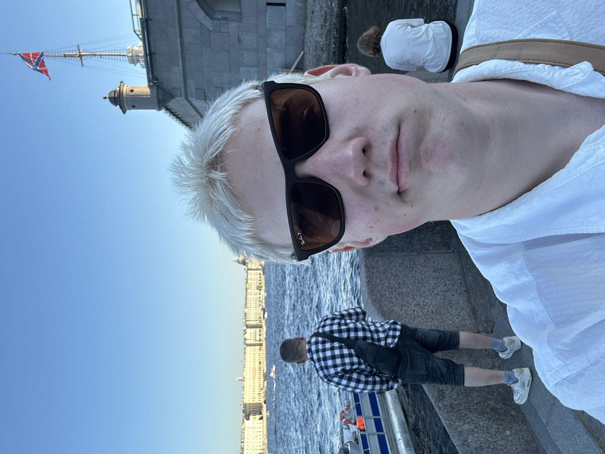 A close-up selfie captures a young, light-skinned individual with short, spiky platinum blonde hair and dark, rectangular sunglasses (with a visible "Ray-Ban P" logo on the left lens) wearing a white, textured, collared shirt. A light brown bag strap crosses their right shoulder. They have a neutral expression, with some reddish blemishes visible on their lower face.
The person is positioned on a granite embankment overlooking a wide, blue, choppy river. In the midground to the left, a man with dark hair, wearing a black and white checkered shirt and dark denim shorts, with a black sling bag across his back, stands facing the river on a stone staircase. He wears white sneakers and grey socks. A white and blue boat is partially visible next to the staircase. To the right of the selfie subject, a person with reddish-brown, medium-length hair, wearing a white t-shirt, sits on a stone ledge, also facing the water. Several small boats are scattered across the distant river.
In the background across the river, a long, neoclassical-style building, light-colored with numerous windows and columns (consistent with the Winter Palace/Hermitage Museum), stretches along the far bank. A prominent golden dome, likely St. Isaac's Cathedral, is visible in the distant skyline to the right of this building. To the right of the selfie subject and behind the other people, a substantial stone fortress-like structure (part of the Peter and Paul Fortress) is visible, featuring a small, cylindrical turret and a tall mast flying the St. Andrew's flag (Russian naval ensign). The sky above is clear and bright blue.
**Location:** Saint Petersburg, Russia, specifically on an embankment of the Neva River, with the Peter and Paul Fortress visible to the right and the Winter Palace/Hermitage Museum across the river.