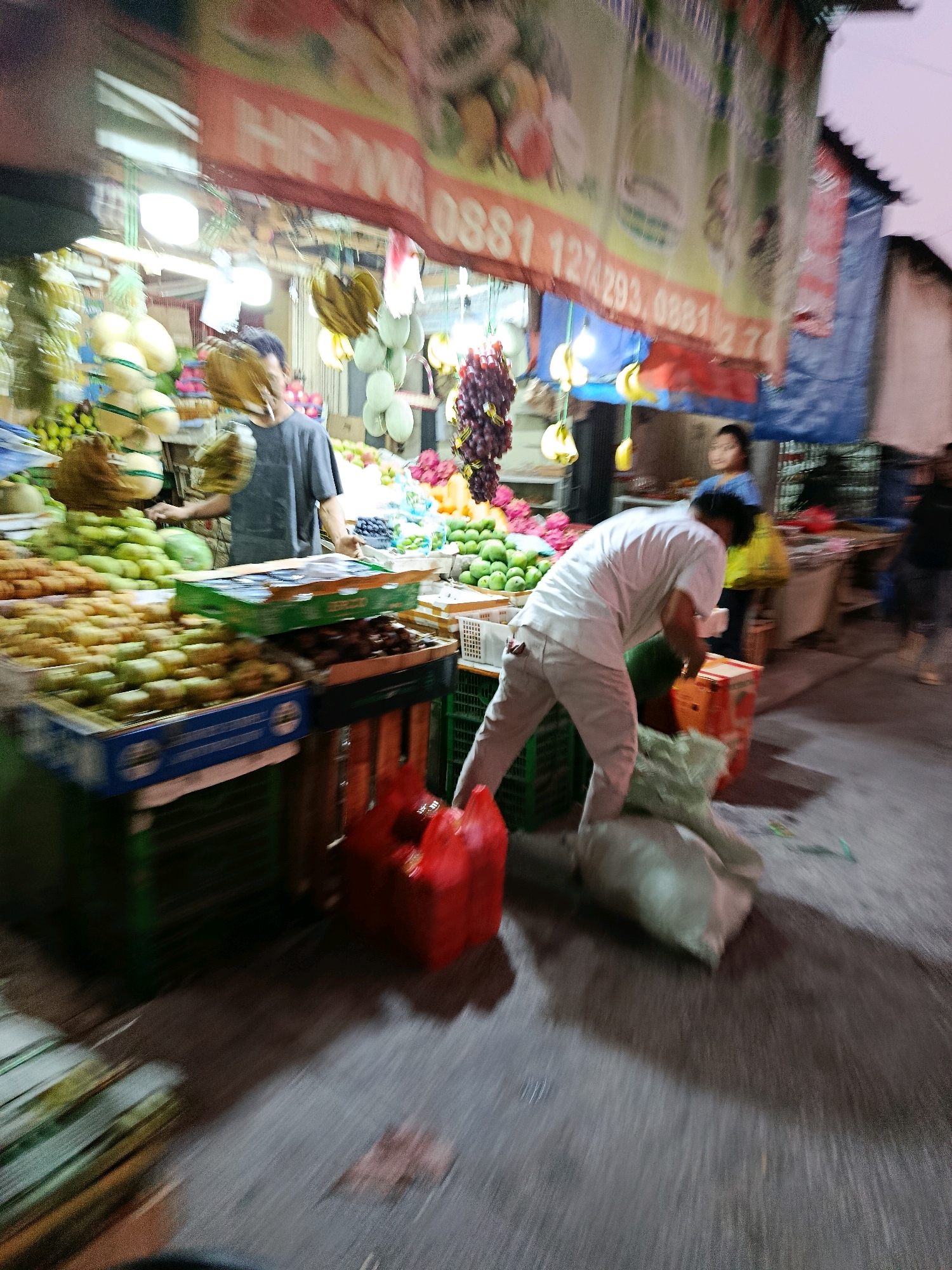 The image depicts an outdoor market scene, likely in Bekasi, Indonesia, at what appears to be dusk or early evening. The market stall is laden with various fruits, including bananas, melons, and what seem to be grapes and durians. A large banner with Indonesian text and what appear to be phone numbers (e.g., "HP/WA 0881...") hangs above the stall.
In the foreground, a man wearing a white long-sleeved shirt and light-colored pants is bending down, appearing to be handling or collecting large sacks. Next to him, a bright red plastic bag is visible. Further back, another individual is partially visible, possibly a customer or vendor, wearing a dark t-shirt. A young woman in a blue shirt is standing behind the fruit stall, looking towards the front.
The ground surface is paved, showing some wet patches and dirt. The lighting is a mix of artificial lights from the stall, casting illuminated areas and shadows, and the ambient twilight. The overall image has a sense of motion blur, suggesting it was taken with a moving camera or during a moment of activity. The atmosphere is typical of a bustling Indonesian market.