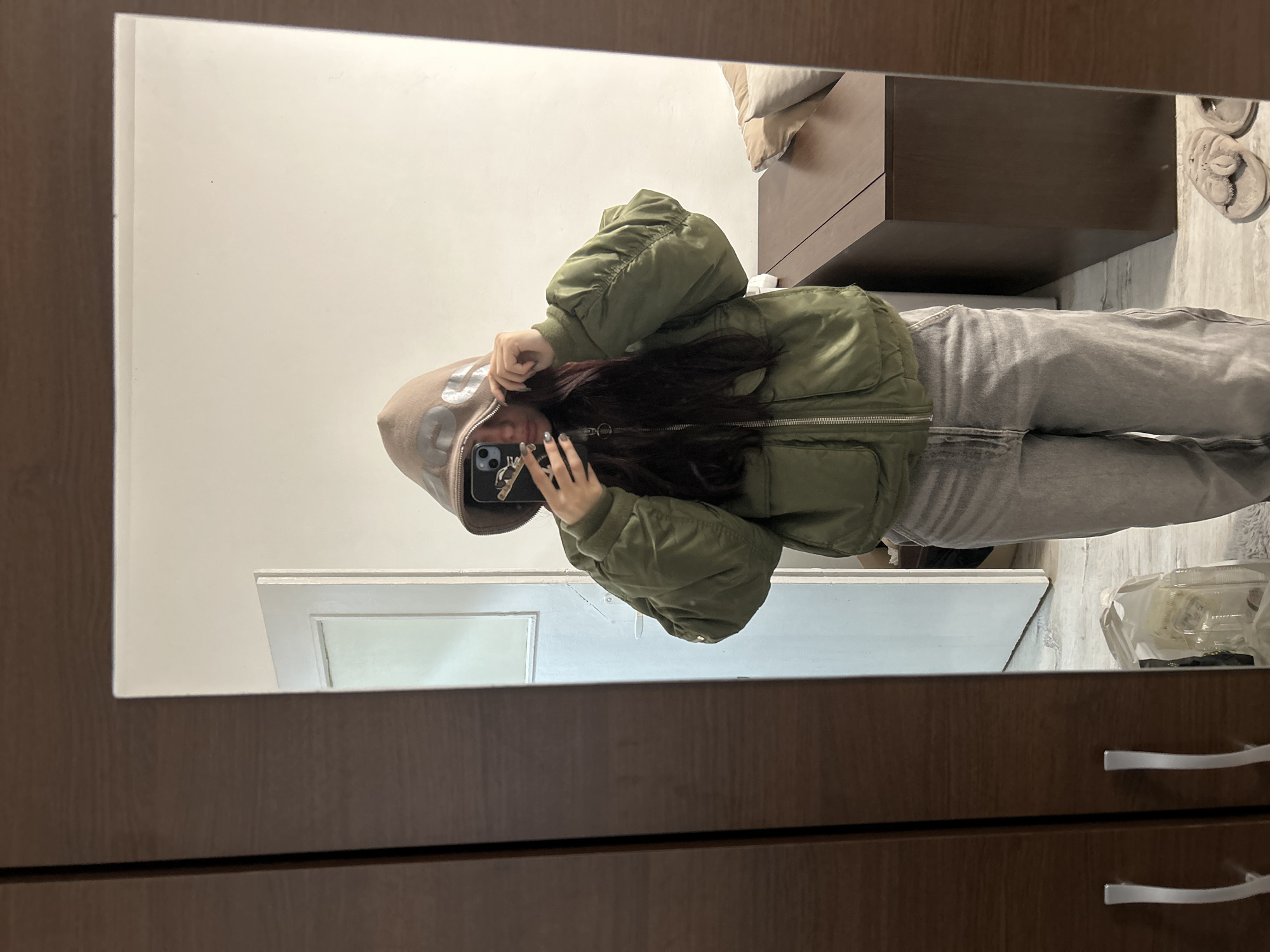 The image shows a person taking a selfie in a mirror. The person is wearing a tan hoodie and an olive green bomber jacket, along with grey pants. They are holding a phone with a case on the back. The reflection shows a portion of a room, including a side table with a pillow, a white door or trim, and a portion of flooring with items on it. The foreground is dominated by the wooden frame of the mirror. There is no information provided that would identify the location as Sofia, Bulgaria.