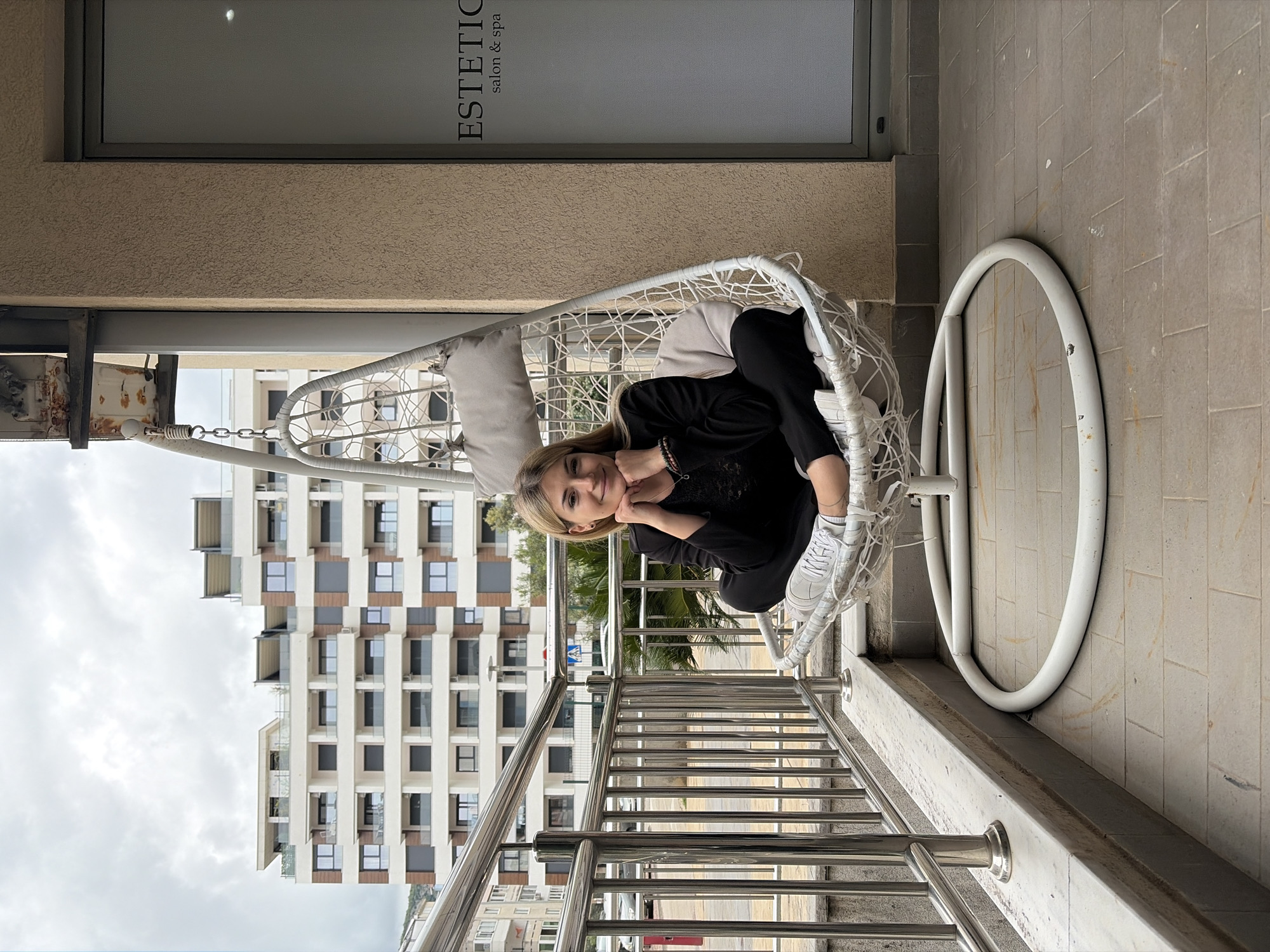 A woman is sitting in a white, egg-shaped hanging chair on a balcony overlooking a street. She is wearing black clothing and white sneakers, resting her face on her hands. The balcony has a stainless steel railing. Behind the railing is a building with a modern facade of white and brown rectangular sections. The sky is overcast. To the right of the chair is a large white circular frame on the tiled floor. Above the balcony is a window with the word "ESTETICA salon & spa." The scene appears to be located in the city of Budva, Montenegro.