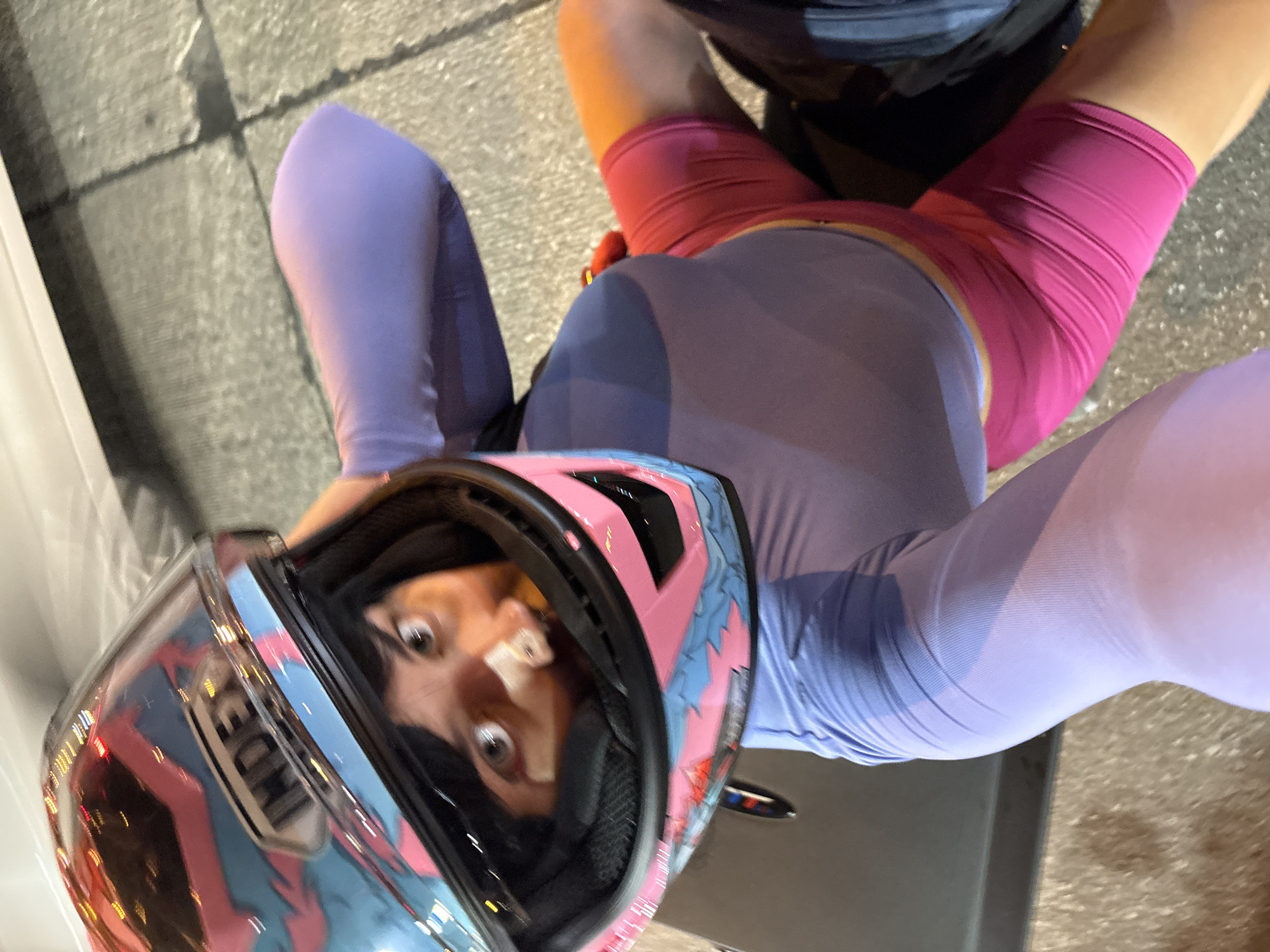 This is a vertical, low-angle, outdoor, daytime photo featuring a person wearing a motorcycle helmet and athletic wear. The person is lying down or sitting, with their legs extended upwards. They are wearing a purple long-sleeved shirt or bodysuit and pink shorts. The helmet is pink with blue and white graphics. The person's face is visible through the helmet's visor, with wide eyes and a surprised expression. There are bandages on their nose.
The background consists of a gray, textured surface, possibly pavement or a concrete walkway. To the left, a white vertical surface, possibly a wall or a car, is visible. The scene appears to be in a residential area, given the address context of "บ้านกาญจน์กนก 23, Thailand". The lighting suggests it is daytime, possibly late afternoon or early evening, given the warm tones in some areas of the image. The overall mood is somewhat playful or dramatic, due to the person's expression and the unconventional pose.
There is visible text on the helmet, specifically the brand name "ICON".