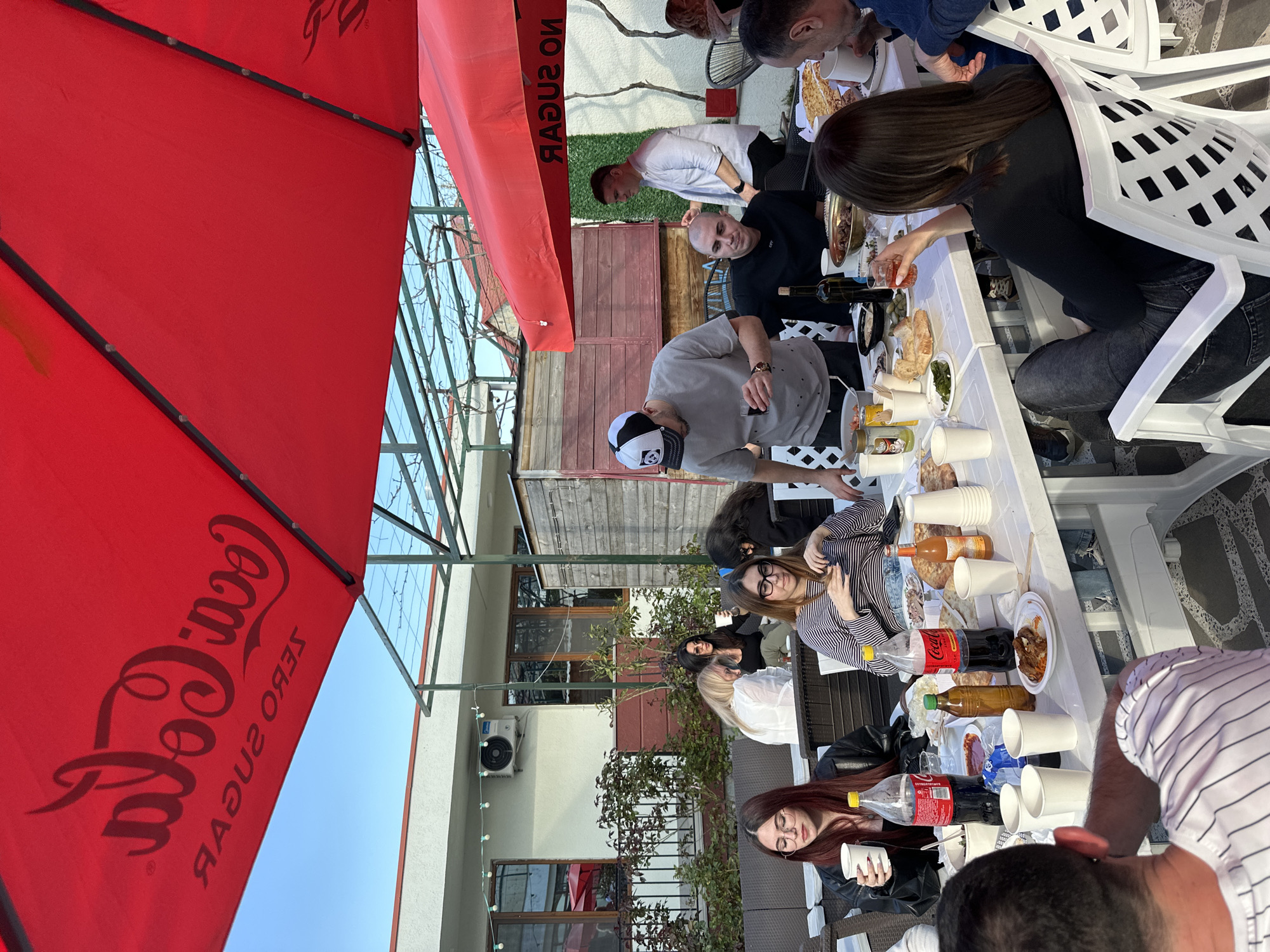 This image captures an outdoor social gathering in Tbilisi, Georgia, taking place on a bright, sunny day. The scene is set on what appears to be a patio or terrace, featuring several white plastic tables arranged for a meal.
**Subjects:** A diverse group of people, including men and women of various ages, are present. Some are seated around the tables on white plastic chairs, while others are standing and interacting. The tables are filled with an array of food and drinks, including large flatbreads, bowls of olives, salads, meat dishes, and numerous white disposable cups. Bottles of Coca-Cola (both regular and Zero Sugar), wine, and other beverages are also visible. A large red Coca-Cola umbrella provides shade over a significant portion of the scene. In the background, there are multi-story buildings with white and wooden facades, interspersed with some greenery and an air conditioning unit.
**Activity:** The individuals are engaged in eating, drinking, and socializing. Several people are reaching for food, pouring drinks, or conversing. The atmosphere appears relaxed and convivial, typical of a casual meal or celebration.
**Notable Details:** The clear blue sky and strong daylight suggest pleasant weather suitable for an outdoor event. The event seems to be a daytime gathering. The layout of the tables and the variety of food indicate a substantial meal.
**Visible Text:** The most prominent text is "Coca-Cola Zero Sugar" (reversed due to the umbrella's printing/angle) displayed on the large red umbrella in the foreground. Another piece of red fabric, possibly a part of another umbrella or banner, also shows "NO SUGAR" in reversed text.