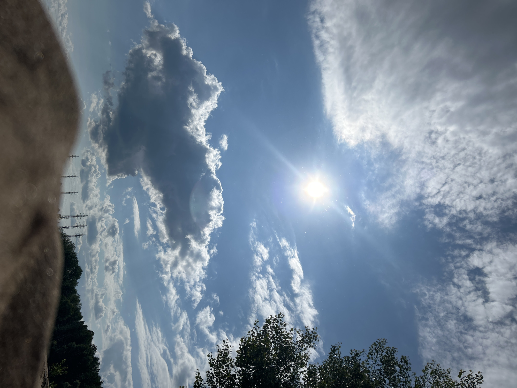 The media file is a photograph of a sky with clouds and the sun. The sun is bright and positioned in the upper center of the image, with a lens flare extending to the left. There are various types of clouds present, from dark, fluffy cumulus clouds on the left to wispy cirrus clouds stretching across the sky. The sky is a clear blue.
In the lower part of the image, there are treetops, suggesting an outdoor setting. On the far left, a blurry, out-of-focus object dominates the foreground, possibly a hand or a textured surface. In the distance, silhouetted against the sky on the left side, are several tall structures that appear to be power line pylons.
The image appears to have been taken during the day, given the brightness of the sun and the clear blue sky. The presence of both dark and light clouds might indicate changing weather or a dynamic sky. The overall impression is one of a bright, possibly warm day with dramatic cloud formations. There is no visible text in the image. The location context is Noviy, Russia.