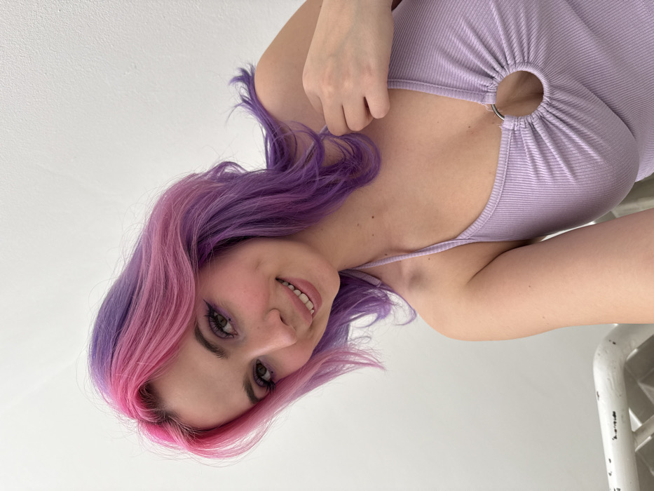 A young female is depicted in a close-up, chest-up portrait, looking directly at the camera with a subtle smile revealing her teeth. She has shoulder-length, wavy hair, dyed pink on the top and front sections, transitioning to purple on the sides and back. Her eyes are accentuated with eyeliner and other makeup, and her lips have a light color. She wears a light lavender, ribbed knit halter top featuring a metallic O-ring detail at the center of the chest, gathering the fabric. Her right hand (viewer's left) gently holds the left strap of her top near her collarbone. The background is a plain, light-colored wall. A blurry, horizontal, light-colored structure, possibly a railing, is partially visible in the lower left foreground. The specific location within the City of Zagreb, Croatia, cannot be determined from the visual information presented in the image.