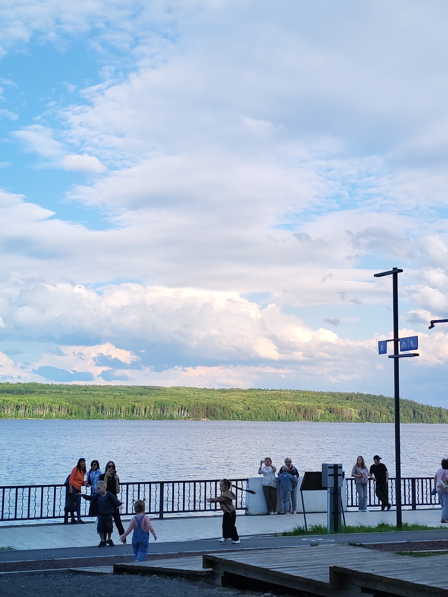 This image captures a scenic view of the Kama River in Chaikovsky, Russia, under a partly cloudy sky. A line of trees and foliage borders the distant shore. In the foreground, a paved walkway runs alongside a black metal railing that overlooks the river. Several people are gathered near the railing, some appearing to take photos. Children are present on the walkway, with one child in blue overalls and another in a brown top and black pants, seemingly playing or running. A lamppost with pedestrian and bicycle signs is visible on the right side of the image, near the walkway. The overall scene suggests a public area, possibly a promenade or park, enjoying the waterfront.