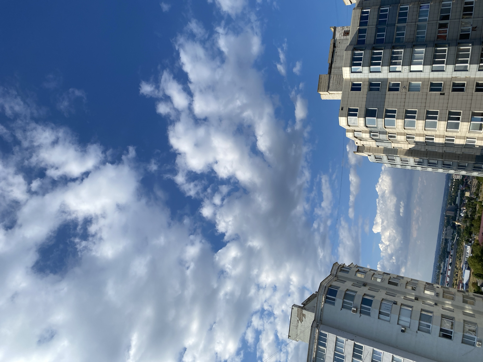 The image captures a view of two apartment buildings against a bright blue sky filled with scattered white and gray clouds. The building on the left is tall and light-colored, with many windows and balconies. The building on the right is also tall and appears to be constructed with a mix of light gray and blue paneling, featuring numerous windows. Both buildings are residential and appear to be in a city environment, with hints of other buildings, greenery, and a distant cityscape visible in the background below the apartment blocks. This scene is located in Molodizhne, Ukraine. There are no people or any obvious actions taking place in the image; it is a static architectural and environmental scene.