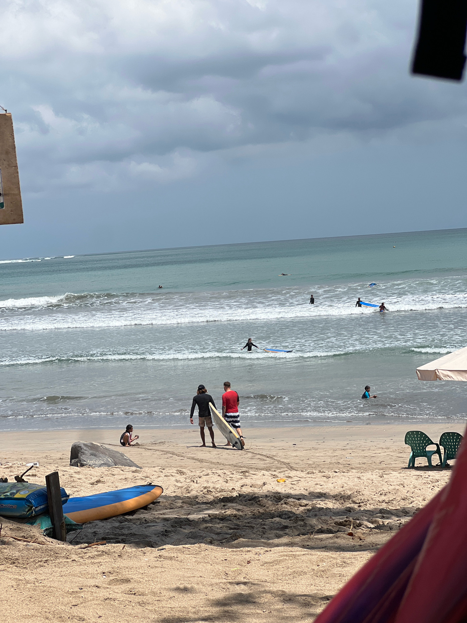 In Kuta, Indonesia, on a beach with sand and the ocean, several people are engaged in or preparing for water activities. Two men stand on the sand near the water's edge, one holding a surfboard. Several other individuals are in the water, some on surfboards and others swimming. The ocean exhibits moderate waves, and the sky is overcast with dark clouds. In the foreground, there are surfboards lying on the sand, along with two empty green chairs.