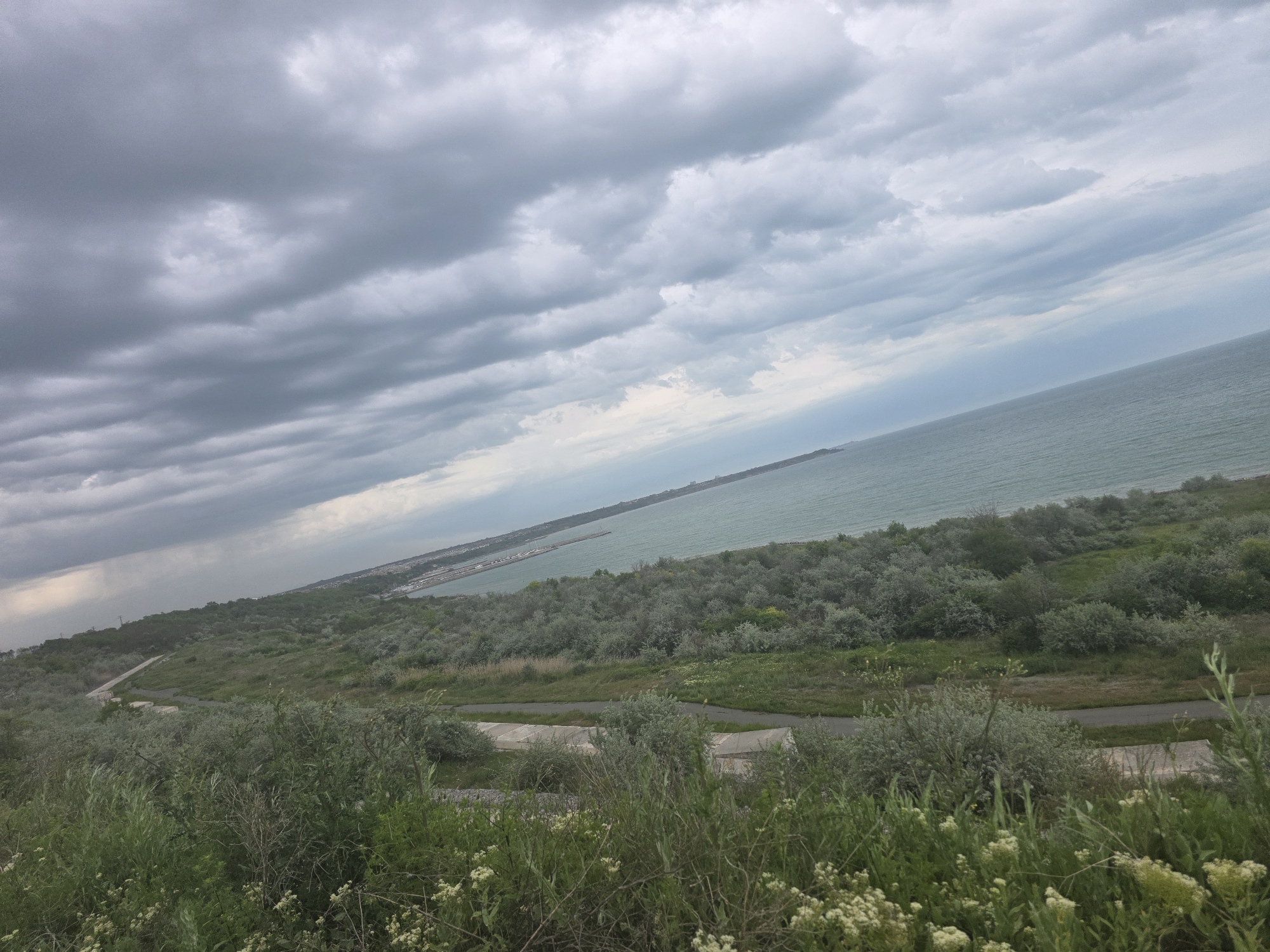 The image shows a view of the coast in Burgas, Bulgaria, under a cloudy sky. The foreground is filled with green vegetation and low-lying shrubs. A road winds through the grassy terrain. In the distance, the coastline stretches along the sea, with buildings and structures visible near the shore. The sea itself is calm. The sky is dominated by grey, overcast clouds.