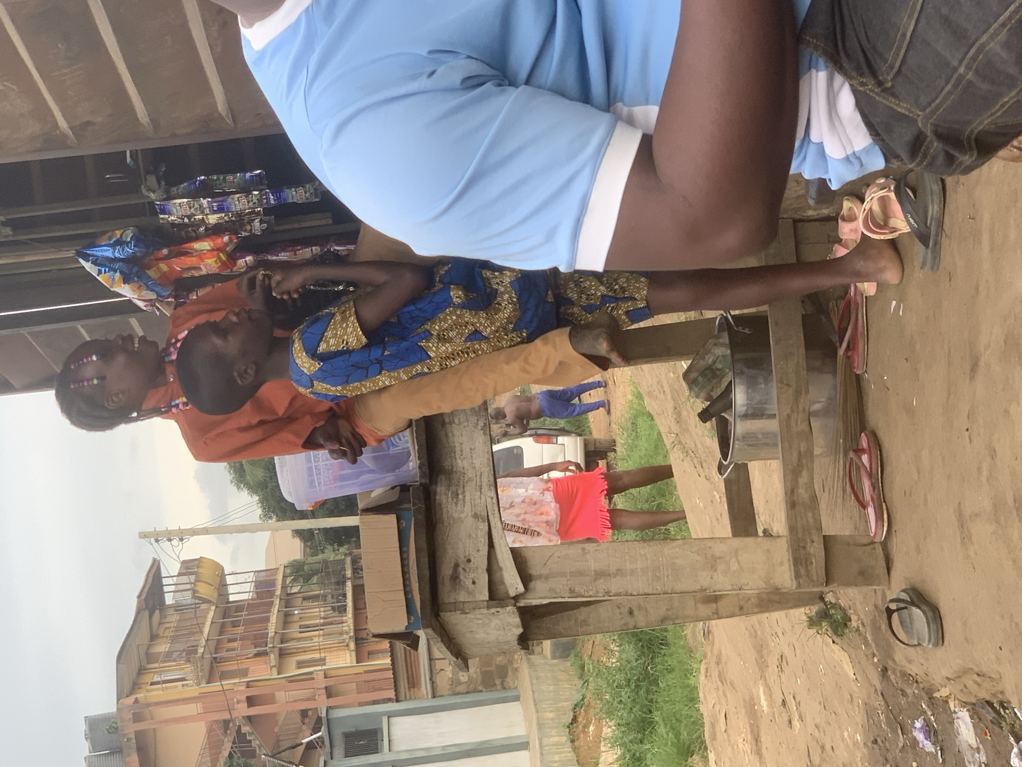 The image depicts an outdoor scene in Shagamu, Nigeria. Two young individuals, a girl with braided hair adorned with colorful beads and a boy wearing a blue and gold patterned garment, are standing behind a rustic wooden table. The girl is wearing an orange top, and the boy's hair is short and dark. The table is laden with a clear plastic container and a cardboard box. Behind them, a collection of packaged goods, possibly snacks or toiletries, hangs from a display. In the background, a multi-story building with balconies and staircases is visible. To the left of the table, a woman in a light-colored top and a vibrant pink skirt is partially visible. Further in the background, a white van and a shirtless man in blue pants are present. The ground appears to be dirt, with patches of green grass and scattered debris. Several pairs of flip-flops are strewn on the ground near the table, including a red pair and a dark grey pair. A metal bucket is also situated beneath the table. The lighting suggests it is daytime, possibly overcast.