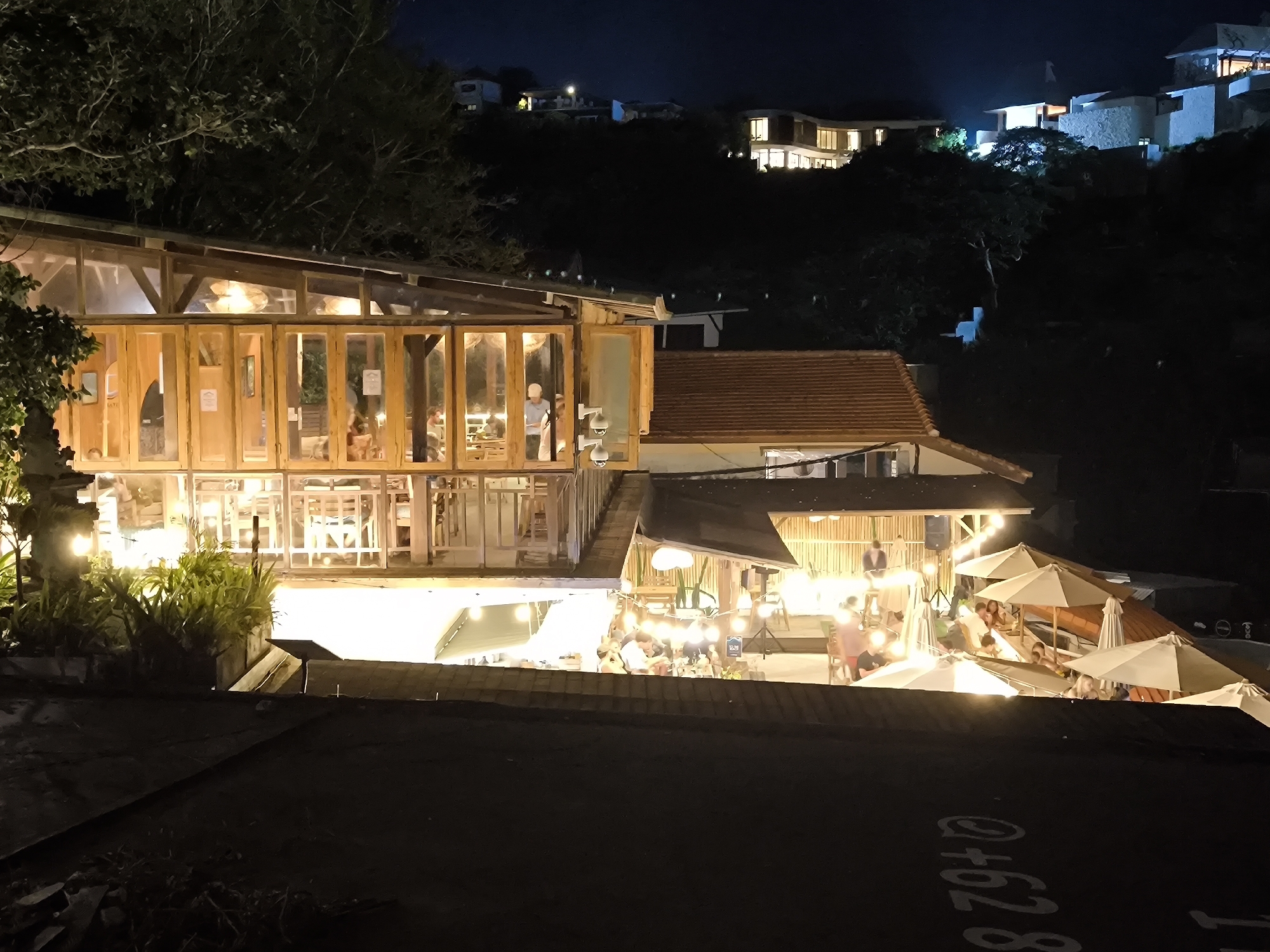 The image shows an outdoor dining and entertainment establishment in Uluwatu, Indonesia, at night. The foreground is dark, showing a rooftop. Above this, a multi-tiered structure houses a restaurant and bar. The restaurant features a glass-walled dining area with wooden frames where people are seated and eating. Next to the restaurant, an open-air bar area has several tables and chairs, lit by string lights. People are dining and socializing in this space. Several white parasols provide shade over the tables. In the background, houses are visible on a hillside, illuminated with lights. The dark sky suggests it is late in the evening. The overall atmosphere is relaxed and inviting.