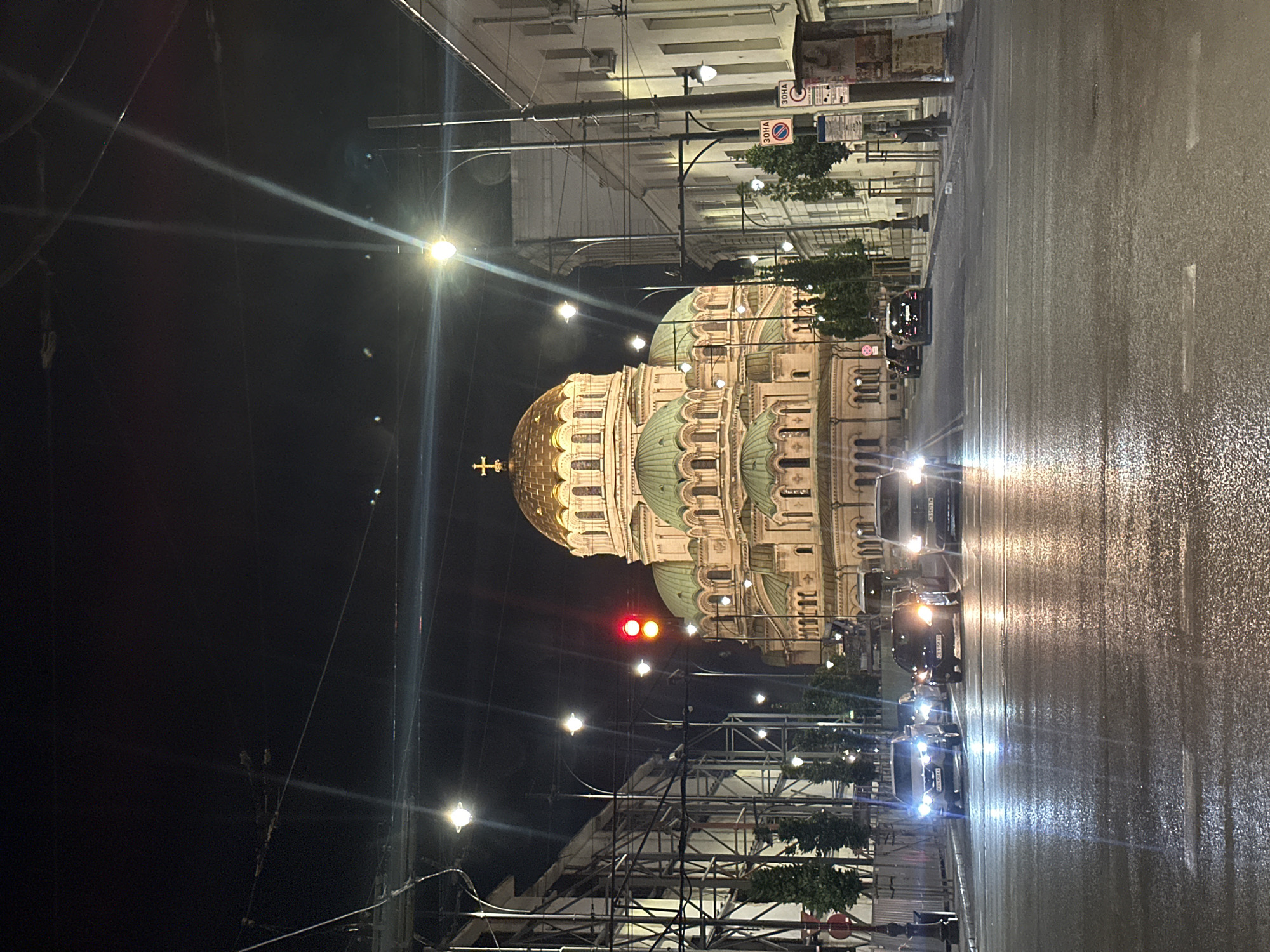 At night, a view shows the Alexander Nevsky Cathedral in Sofia, Bulgaria. The cathedral’s golden dome is visible, along with several green domes. There are street lights along the street and construction scaffolding. The street looks wet and reflects the lights of moving vehicles. A red traffic signal is visible. Buildings are visible on both sides of the street. The sky is dark.