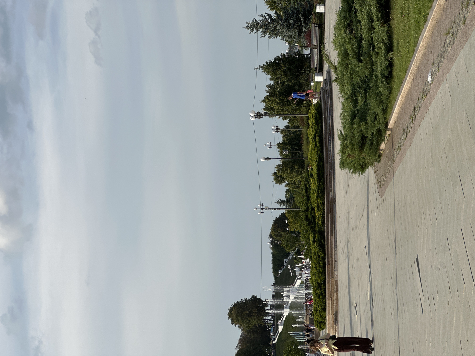 The image depicts a lively public park scene, likely in Rusanovo, Russia, on a bright, possibly late afternoon given the long shadows. The sky is a pale, overcast blue. The foreground features a wide, paved walkway that curves to the right, bordered by lush, green bushes and manicured hedges. Further into the scene, a long row of lampposts illuminates a tree-lined promenade. Beyond the trees, a series of fountains are active, with water spraying upwards, suggesting a warm day or a planned water display. Several people are visible, some walking along the path, others seated, and a few interacting near the fountains. One individual in the foreground is on a scooter. The overall atmosphere appears relaxed and leisurely, with people enjoying the outdoor amenities. The presence of benches amidst the greenery and the general layout of the park suggest a well-maintained urban recreational space.