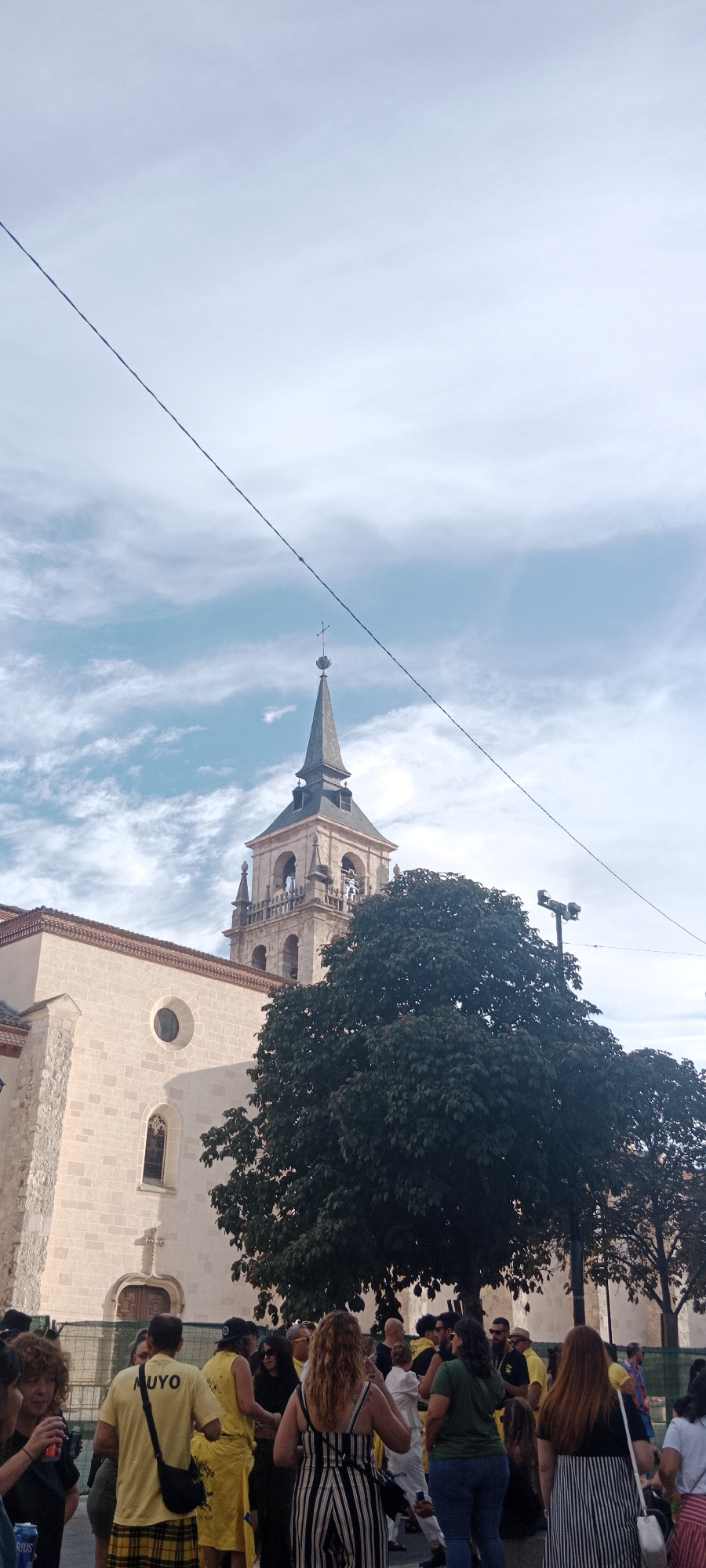 In Alcalá de Henares, Spain, a crowd gathers in front of a stone church with a prominent bell tower. The tower features arched openings and a spire topped with a cross. A large, dark green tree partially obscures the view of the church's side and the bell tower. In the foreground, a diverse group of people are visible, some wearing yellow shirts and plaid skirts, others in striped outfits. Some individuals are holding drinks and engaging in conversation. The sky above is a pale blue with scattered white clouds, and dark wires stretch across the upper portion of the frame. The overall scene suggests a public gathering or event taking place outdoors.