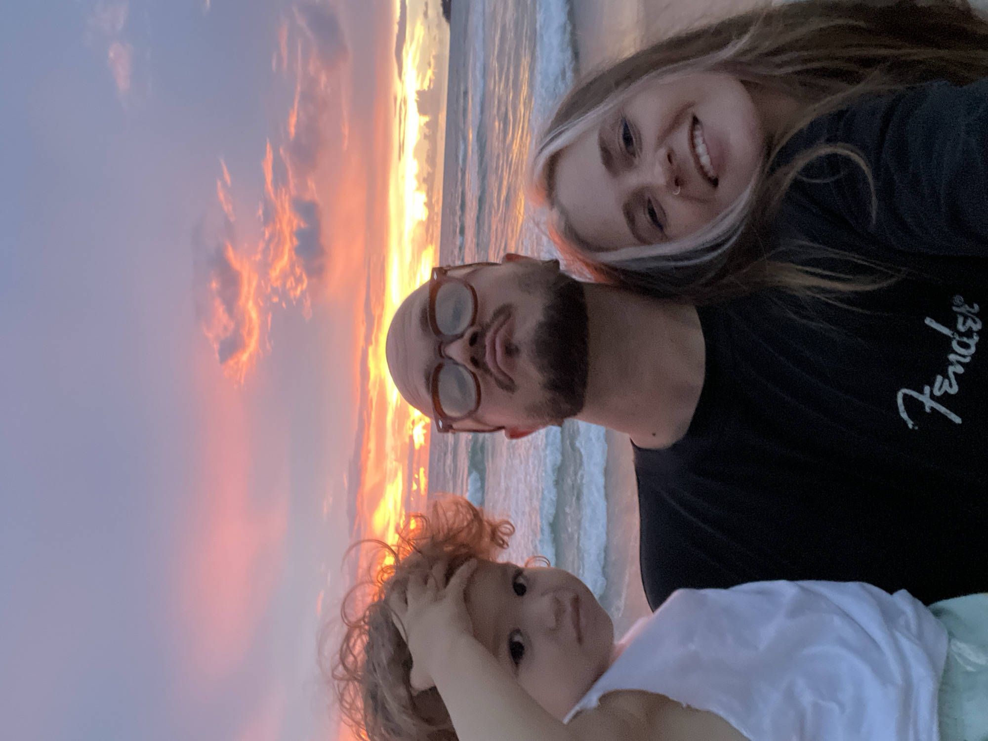 This is a selfie of three people on a beach at sunset. The subjects are two adults and a young child. The adult in the foreground on the right is a woman with blonde hair, smiling at the camera. She is wearing a dark t-shirt with a white logo. The adult in the middle is a man with a beard and glasses, looking towards the woman. He is wearing a dark t-shirt. The child is in the bottom left of the image, looking up with a curious expression.
The background is a beach scene with the ocean and a colorful sunset. The sky is filled with dramatic clouds painted in shades of pink, orange, and yellow, indicating it is either sunrise or sunset. The ocean has small waves rolling onto the shore.
The overall emotion of the image is joyful and familial, with the people appearing relaxed and happy. The time of day is clearly sunset due to the lighting and colors of the sky. There is no discernible location context beyond it being a beach. The visible text on the man's shirt appears to be a logo, possibly from a brand.