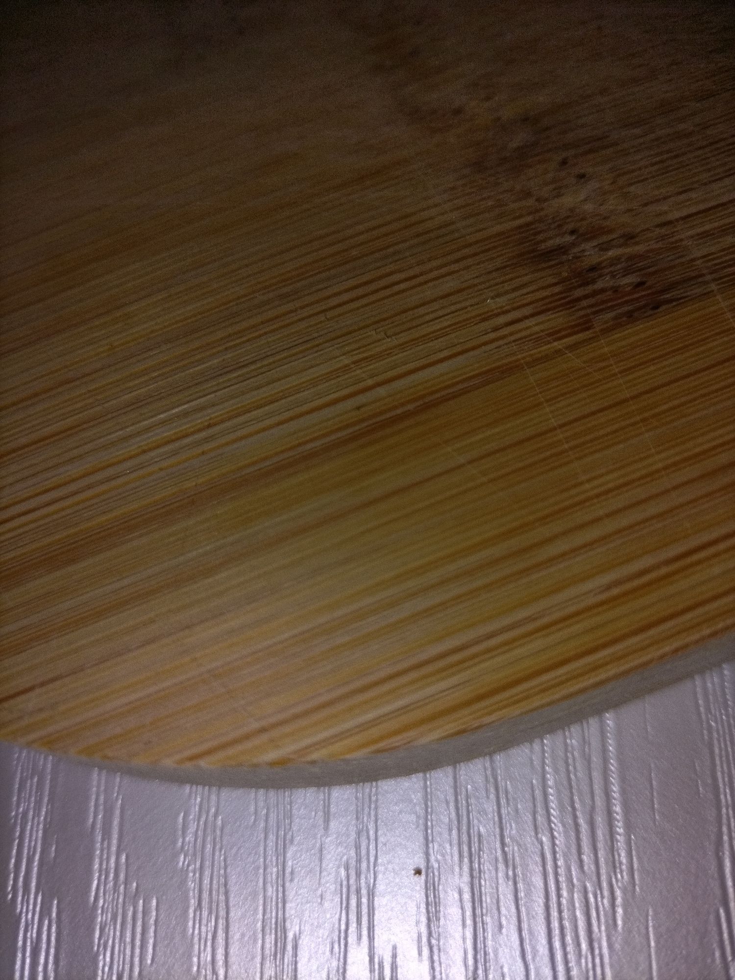 A close-up, overhead shot shows a light-brown, wooden surface, likely a cutting board or tabletop, with a curved edge. The wood grain runs diagonally from the top left to the bottom right of the frame. Darker spots and faint scratches are visible on the wood. Below the wooden surface is a textured, white surface with vertical ridges. The lighting appears to be dim, with the top portion of the image being darker than the bottom.