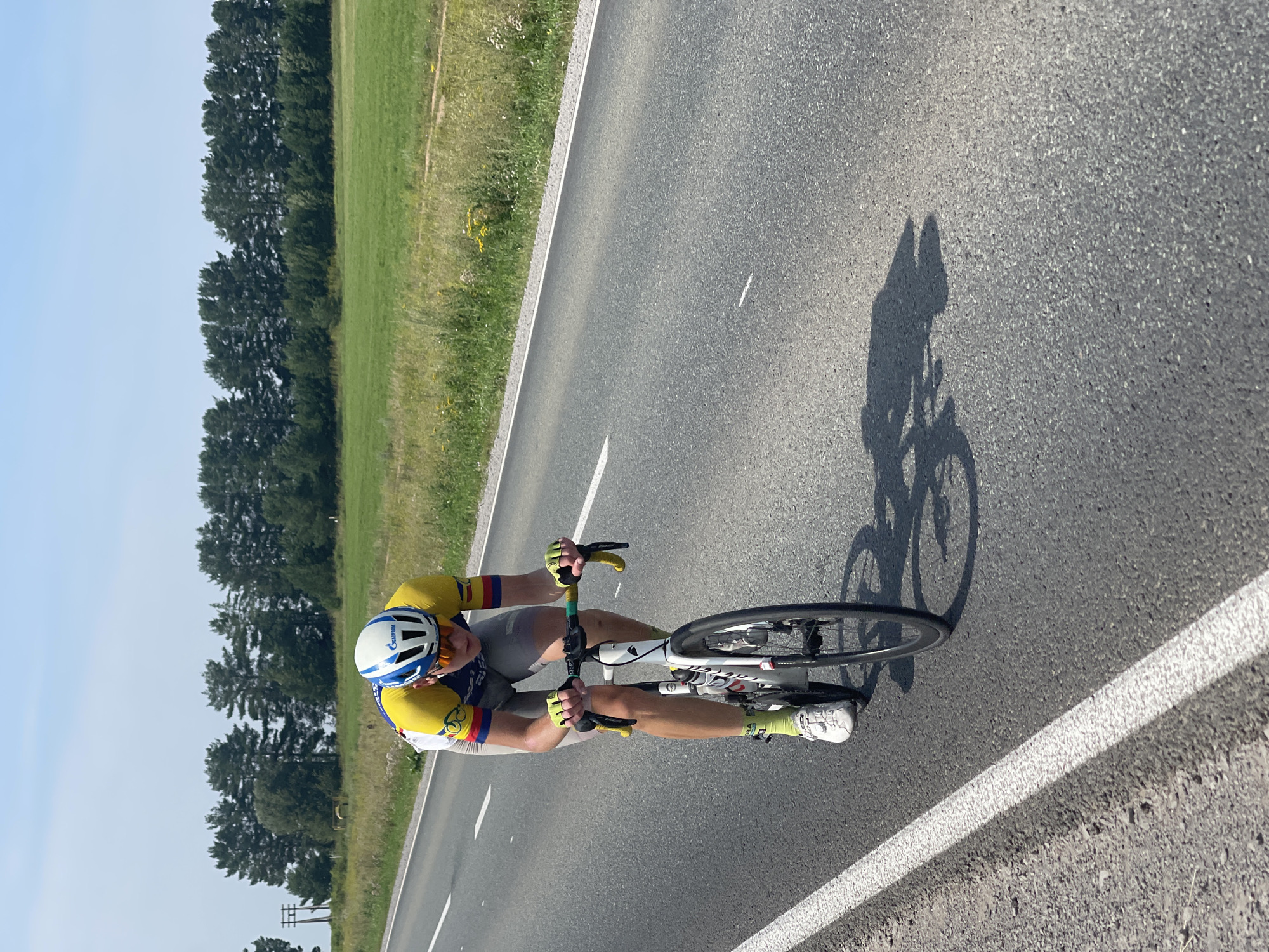 A cyclist in a yellow and blue racing suit and a white helmet with blue accents rides a white racing bicycle down an asphalt road. The cyclist is wearing sunglasses, yellow gloves, and green socks. The road has a white line marking the edge. In the background, there is a field of green grass and a line of trees under a clear blue sky. A shadow of the cyclist and bicycle is cast on the road. The location is Perevoznoe, Russia.