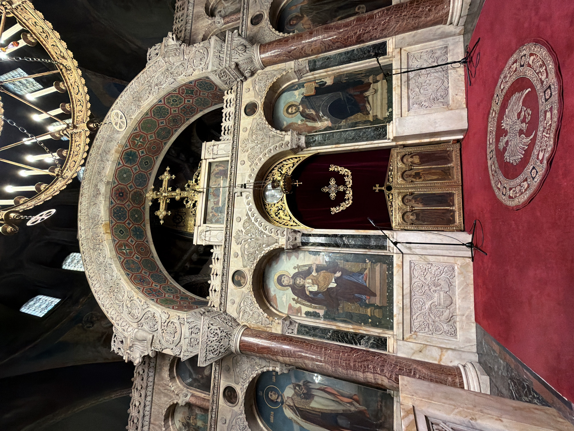 This is an indoor photograph taken within the Alexander Nevsky Cathedral in Sofia, Bulgaria. The image captures a view of the cathedral's interior, focusing on the iconostasis and the area in front of it. The scene is richly decorated with religious iconography and ornate architectural elements.
In the foreground, a vibrant red carpet covers the floor. A circular rug with an intricate design, featuring a double-headed eagle, is prominently placed on the red carpet. Two microphone stands are visible, one to the left and another to the right of the central section.
The iconostasis, a wall of icons, dominates the midground. It features several large, arched panels containing religious paintings, or icons. To the left, an icon depicts a saint in white and gold robes. The central icon shows the Virgin Mary holding the infant Jesus. To the right, an icon portrays Christ holding a book. The doors of the iconostasis are ornate and gilded, flanked by these icons. Above the central doors, a decorative red curtain is hung, adorned with a golden emblem. A golden chandelier hangs from the ceiling above the iconostasis.
The architectural elements are characterized by marble columns with intricately carved capitals. The arches above the icons are adorned with geometric patterns and carved stone work. The ceiling is dark and vast, with some windows visible in the upper portion of the image, allowing natural light to filter through. The overall impression is one of grandeur and spiritual solemnity. There are no people visible in the frame.