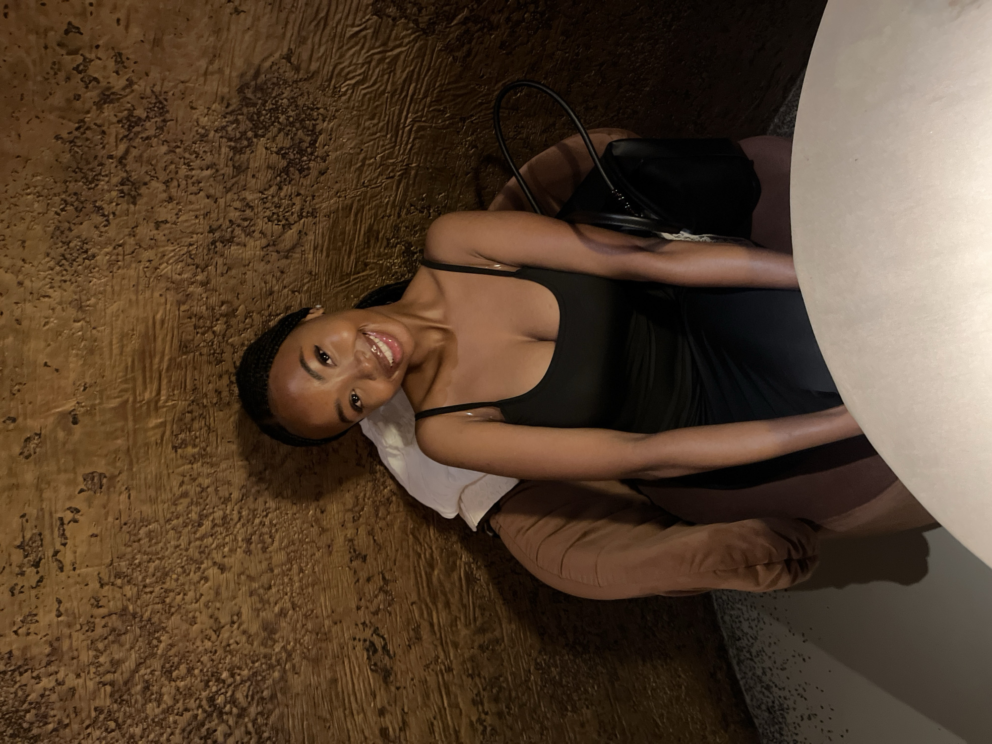A young Black woman with braided hair and a black dress is sitting in a brown chair. She is smiling at the camera. A black purse is on the chair next to her. The background is a textured, brown wall. The woman is in Kazan, Russia.