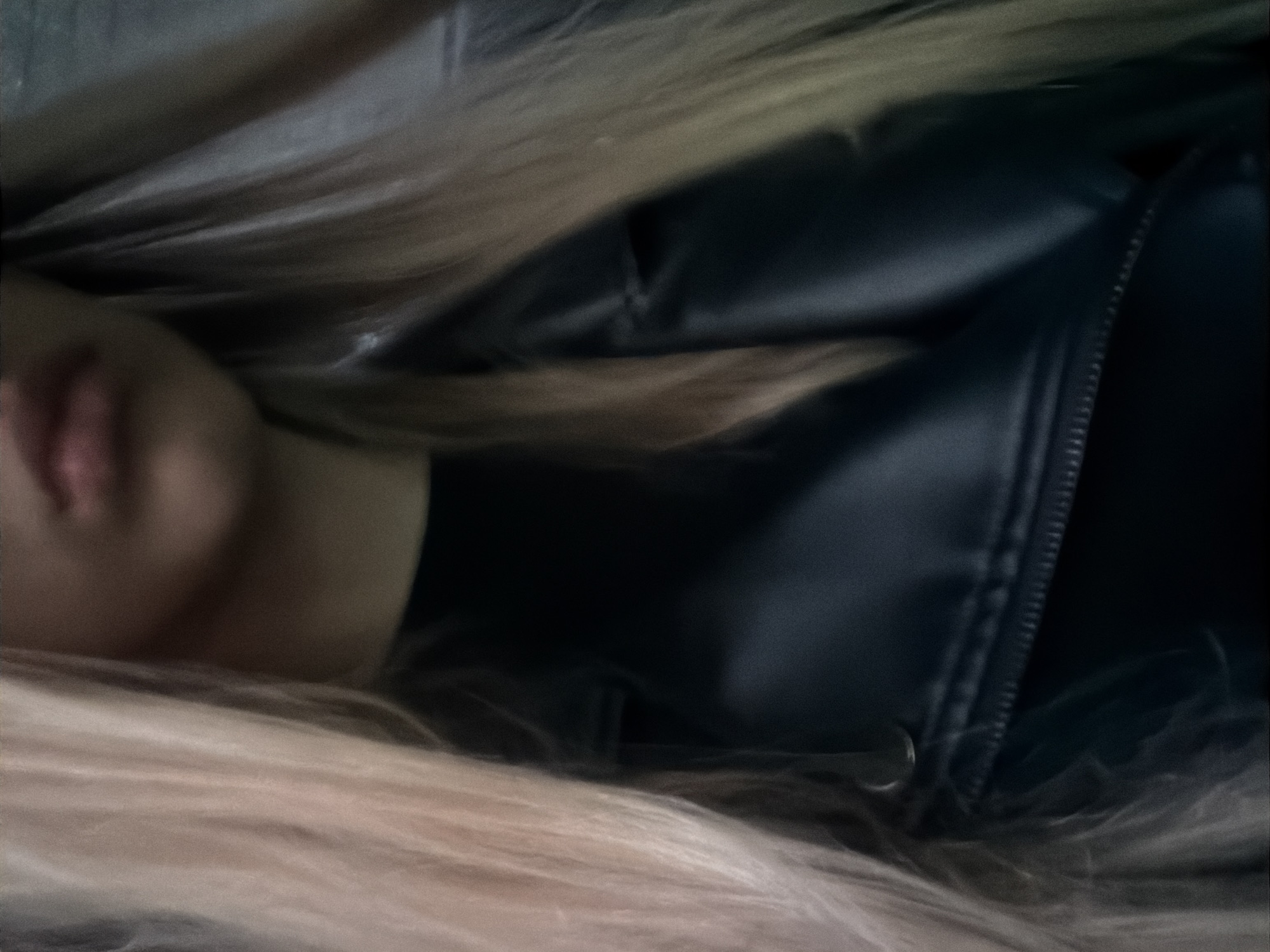 A close-up, tilted shot shows a person lying down, seemingly indoors in Unknown, Unknown. The subject has fair skin, and their lips are visible. Long, light-blonde hair surrounds their face and body. The person is wearing a dark, possibly black leather or faux leather jacket with a visible zipper. The lighting is dim, and the focus is soft, blurring details in the background.