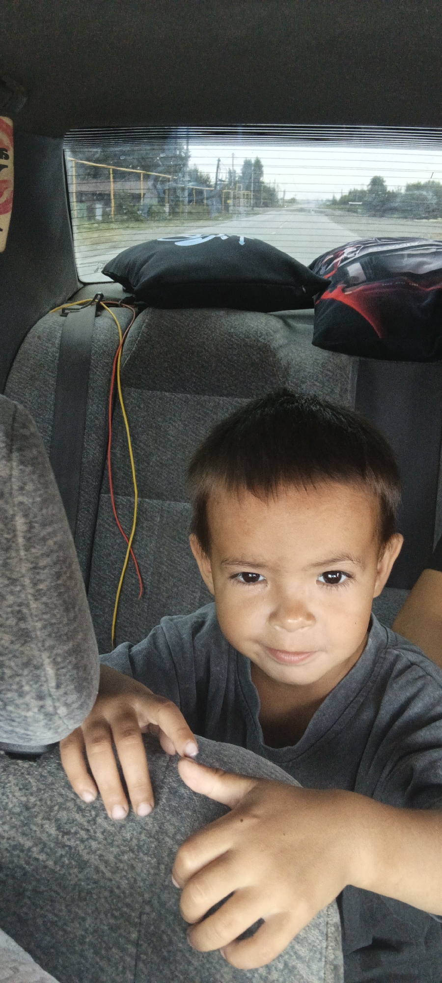 The image depicts a young boy in the back seat of a car, looking towards the camera. The boy has dark hair and is wearing a grey t-shirt. His hands are resting on the seat in front of him. In the background, through the car's rear window, a road can be seen with trees and industrial structures. The car's interior includes a grey seat, a black pillow, and some red and yellow wires. This scene is likely captured within the city of Kargat, Russia, given the context provided.