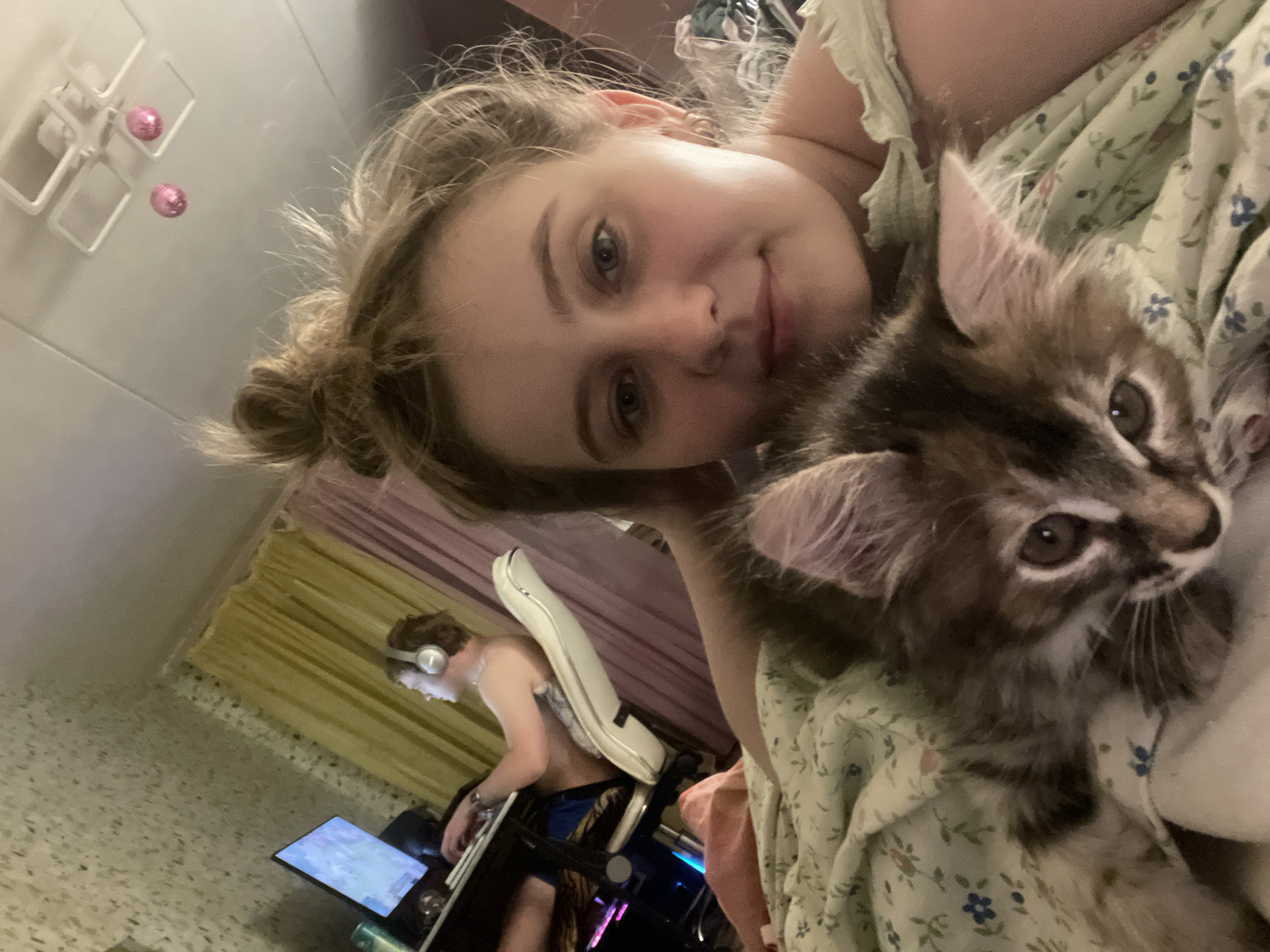 In an indoor scene, a young woman with blonde hair in a messy bun is taking a selfie with a fluffy kitten. She is wearing a light-colored top with a ruffled neckline. The kitten is positioned on her chest, peeking out from under her chin. In the background, a person is sitting in a white gaming chair, wearing headphones and looking at a computer screen. The room appears to be a bedroom, with yellow and purple curtains visible, and a ceiling fixture with pink decorative balls. The overall impression is of a casual, domestic setting.