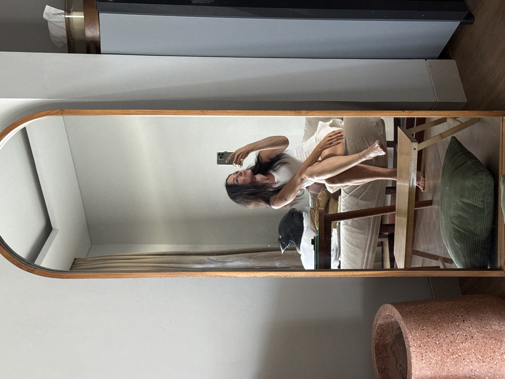 A woman with long dark hair, wearing a white tank top and white skirt, is taking a mirror selfie in a dimly lit room. She is sitting on a sofa, with her right leg crossed over her left. Her left arm is extended, holding a smartphone, and her head is tilted back slightly as she looks at her reflection. A black motorcycle helmet rests on a pillow behind her. In front of the sofa is a low wooden table with a wicker basket on it, and a small wooden stool with a green cushion on the floor. A tall, arched mirror with a wooden frame dominates the center of the image, reflecting the scene. To the left of the mirror is a large, textured pink planter. To the right of the mirror is a tall gray cabinet. The room appears to be a residential space, possibly in North Kuta, Indonesia, with neutral-colored walls and wooden flooring.