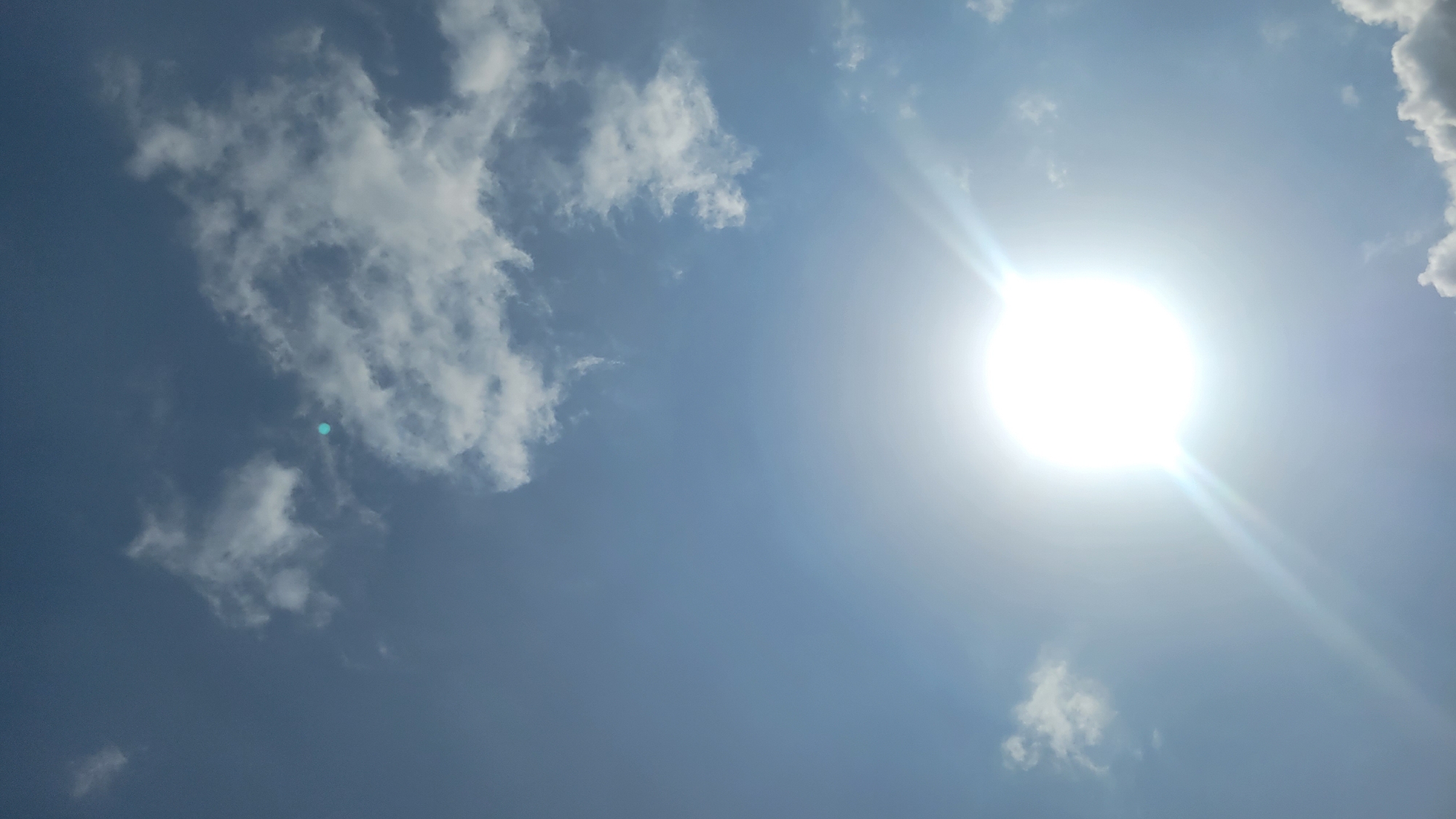 A vertical, full-shot photograph captures the bright sun shining through a clear blue sky with scattered white clouds. The sun is positioned in the lower center of the frame, emitting strong rays that create a lens flare effect. A small, distinct blueish-green circular object is visible in the upper portion of the sky, near a cluster of clouds. There are no people or man-made objects discernible in the image, suggesting an outdoor scene in an open area. The lighting indicates it is daytime.