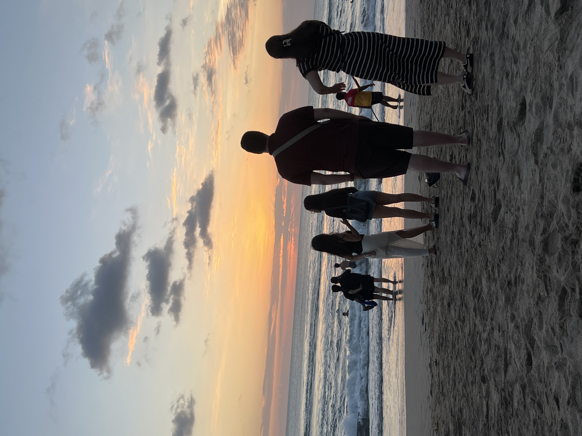 The image depicts a beach scene in Kuta, Indonesia, at sunset. The sky is filled with vibrant orange and pink hues, with scattered clouds. Several people are visible on the sandy shore and in the ocean. In the foreground, a group of four individuals stands facing the water, with two of them in silhouette. To the left, a couple walks into the shallow waves. Further out in the water, other people can be seen swimming or wading. The ocean has moderate waves crashing onto the shore. The sand is textured with footprints and ripples.