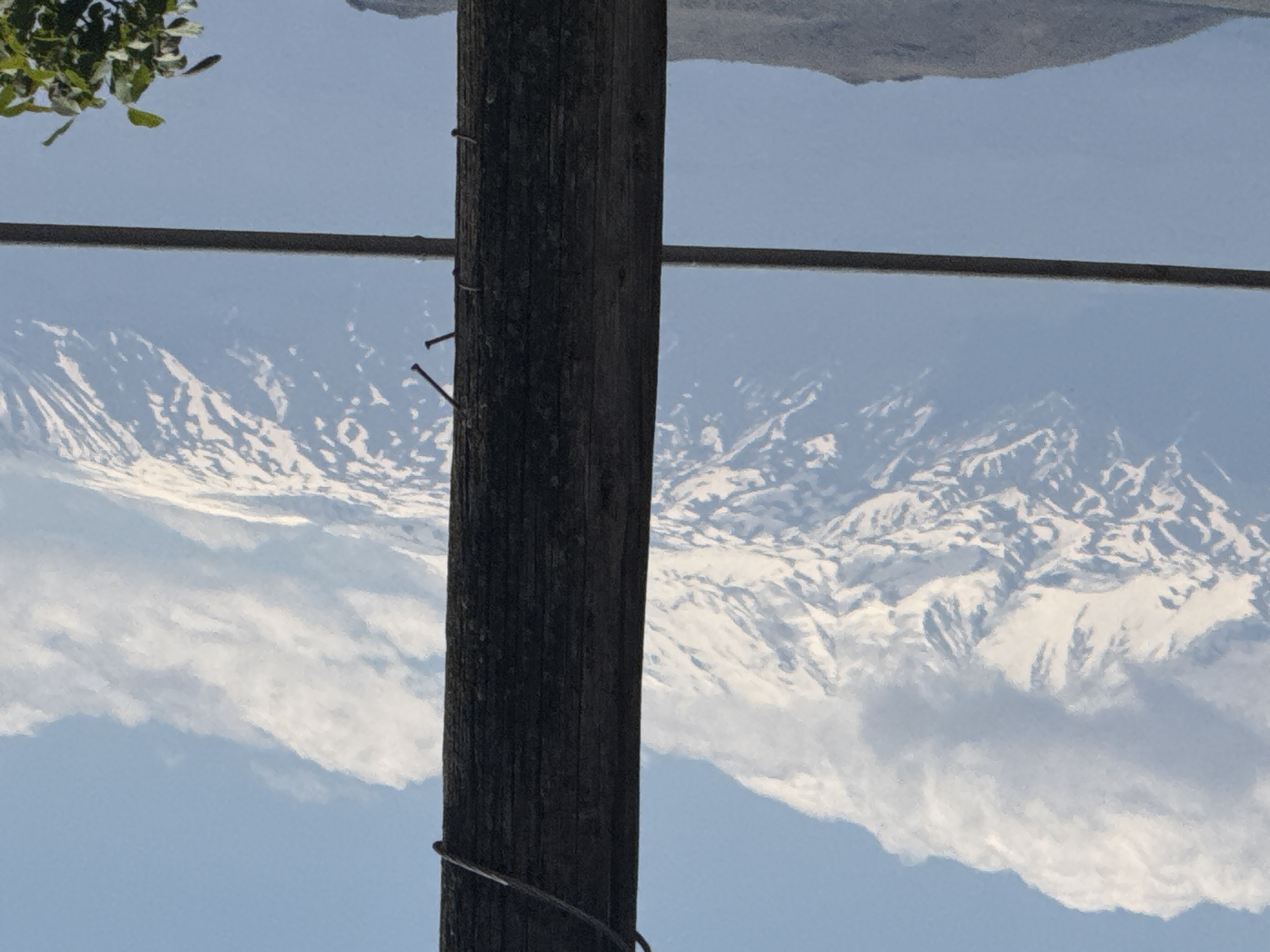 The image is oriented upside down, featuring a wooden utility pole in the foreground, slightly off-center to the left. Three nails are visible protruding from the pole. A thin, dark cable is wrapped around the lower part of the pole. In the background, there is a landscape featuring snow-capped mountains against a light blue sky with clouds. A black cable stretches horizontally across the upper part of the frame. Some green foliage is visible in the upper left corner. The location is Unknown, Unknown.