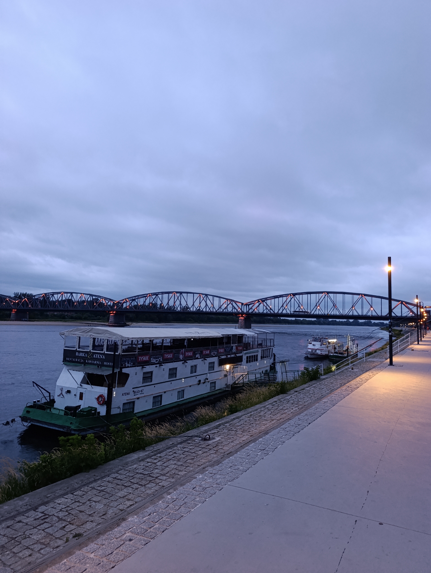 This is a wide-angle, eye-level shot taken at dusk of the Vistula River in Toruń, Poland, looking towards the historic bridge. A large, white barge named "BARKA ATENA" is moored to the left, featuring a restaurant, cafe, and hostelry. The barge has multiple windows and a deck area with seating. A smaller white boat is also visible docked further downriver. The river is calm and reflects the dim light. The foreground is dominated by a paved walkway with cobblestone edging and modern lampposts casting warm light. In the background, the prominent truss bridge, likely the Toruń Rail Bridge, spans the river, its structure illuminated with orange lights. The sky is overcast and a deep twilight blue. There are no people clearly visible in the scene. The overall atmosphere is serene and the lighting suggests the early evening.