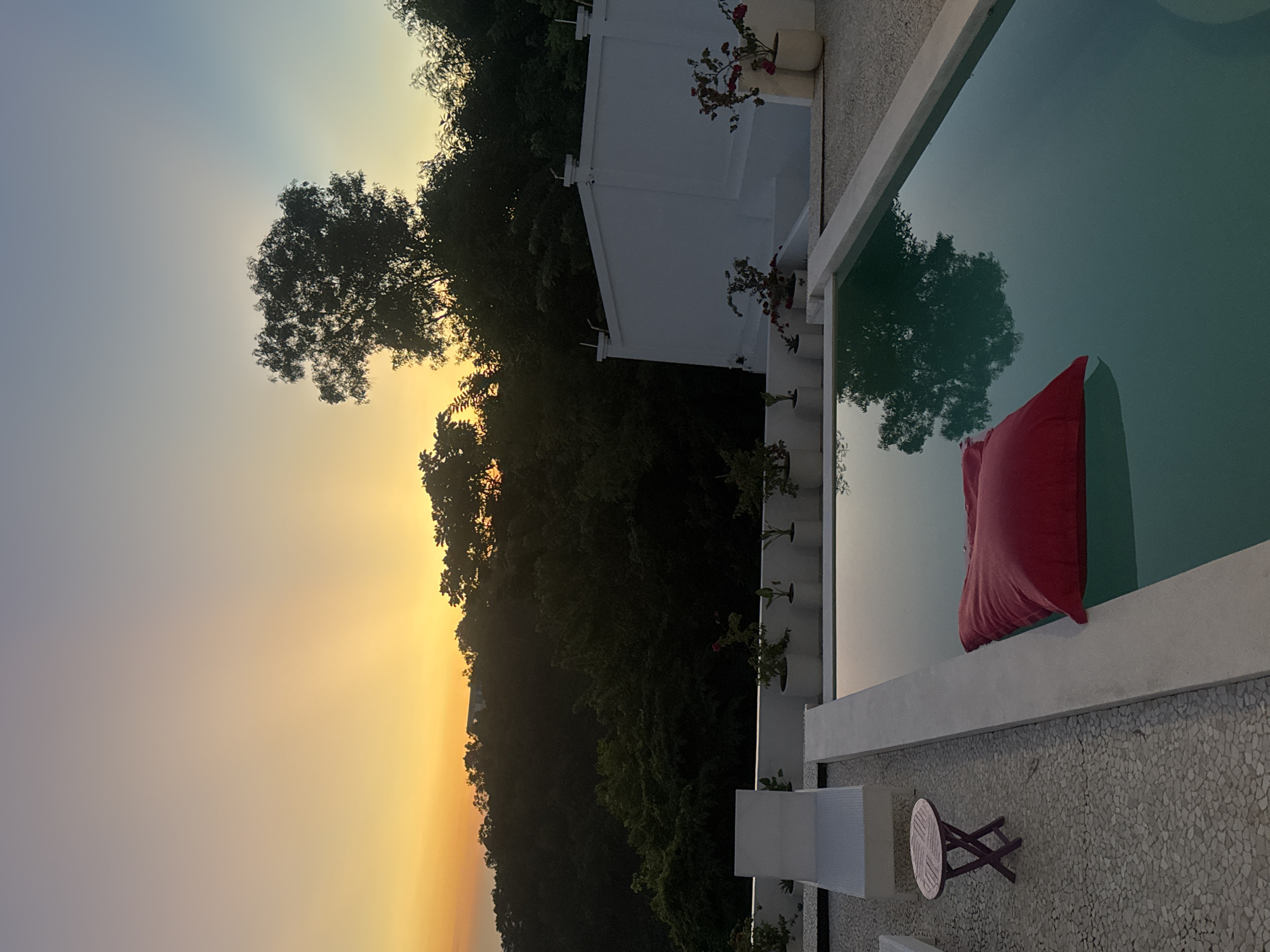 The image captures a tranquil scene in Kuta Selatan, Indonesia at dusk. A rectangular swimming pool occupies the right side, reflecting a tree and the fading light of the sky. A red cushion rests partially submerged within the pool. Adjacent to the pool is a white patio area with a small round wooden table and two white chairs. The patio floor is covered in small pebbles.
Behind the pool, a white building stands adorned with flower pots. Beyond the building, a dense area of trees blankets a hill, silhouetted against the setting sun. The sky transitions from a pale blue at the top to a vivid orange near the horizon.