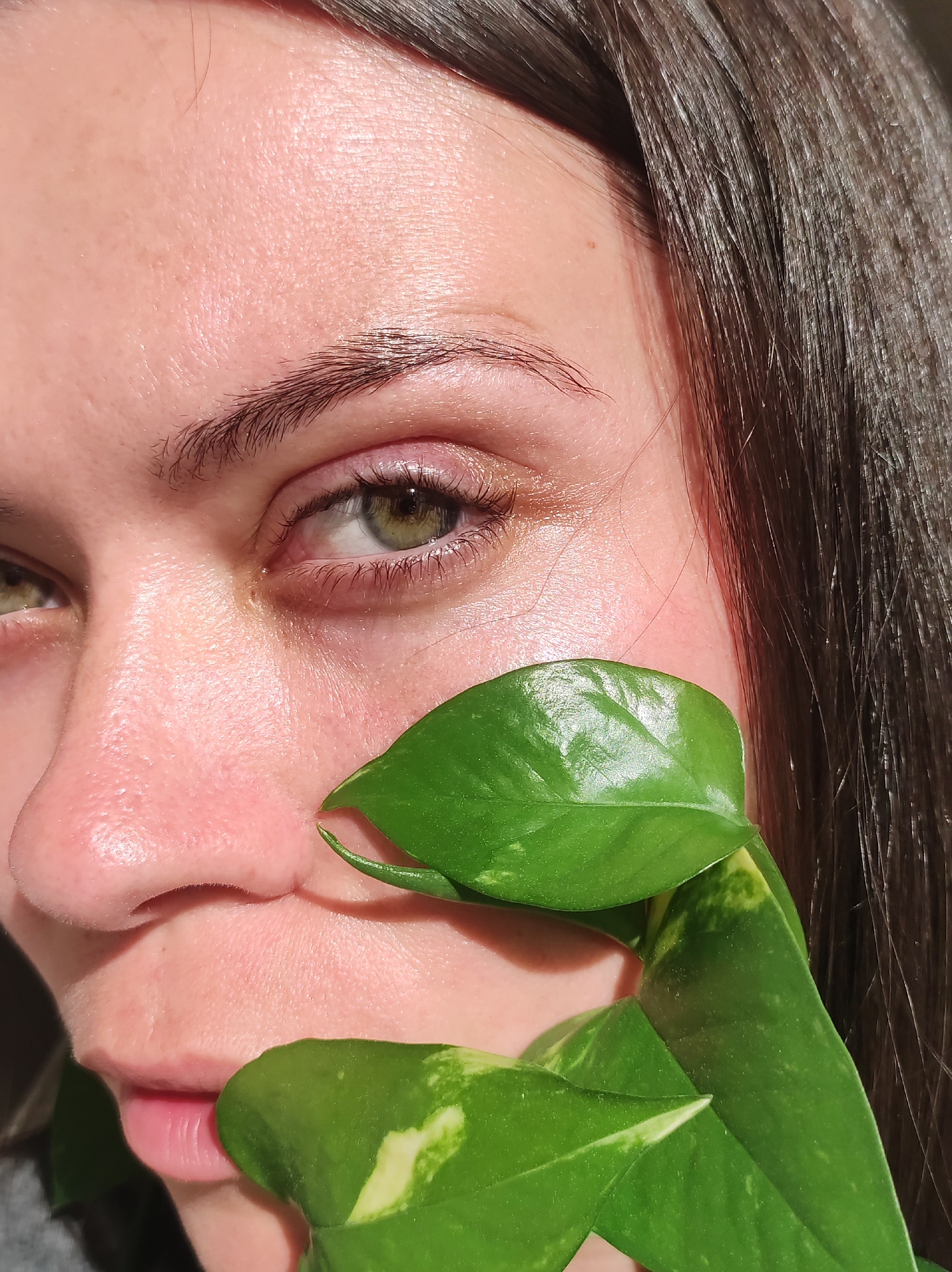 A close-up, eye-level shot focuses on the right side of a person's face, specifically their eye, nose, and lips, partially obscured by a vibrant green leaf of a pothos plant. The individual has fair skin with visible pores and a subtle sheen, suggesting natural light exposure. Their left eye, with a green iris and dark lashes, is looking directly at the camera. The eyebrow above is dark and well-defined. The nose is slightly pink, and the lips are a natural pink hue. The dark brown hair of the individual frames the right side of the image. The background is dark and out of focus, highlighting the subject. There are no discernible landmarks to place this image in Chaikovsky, Russia.